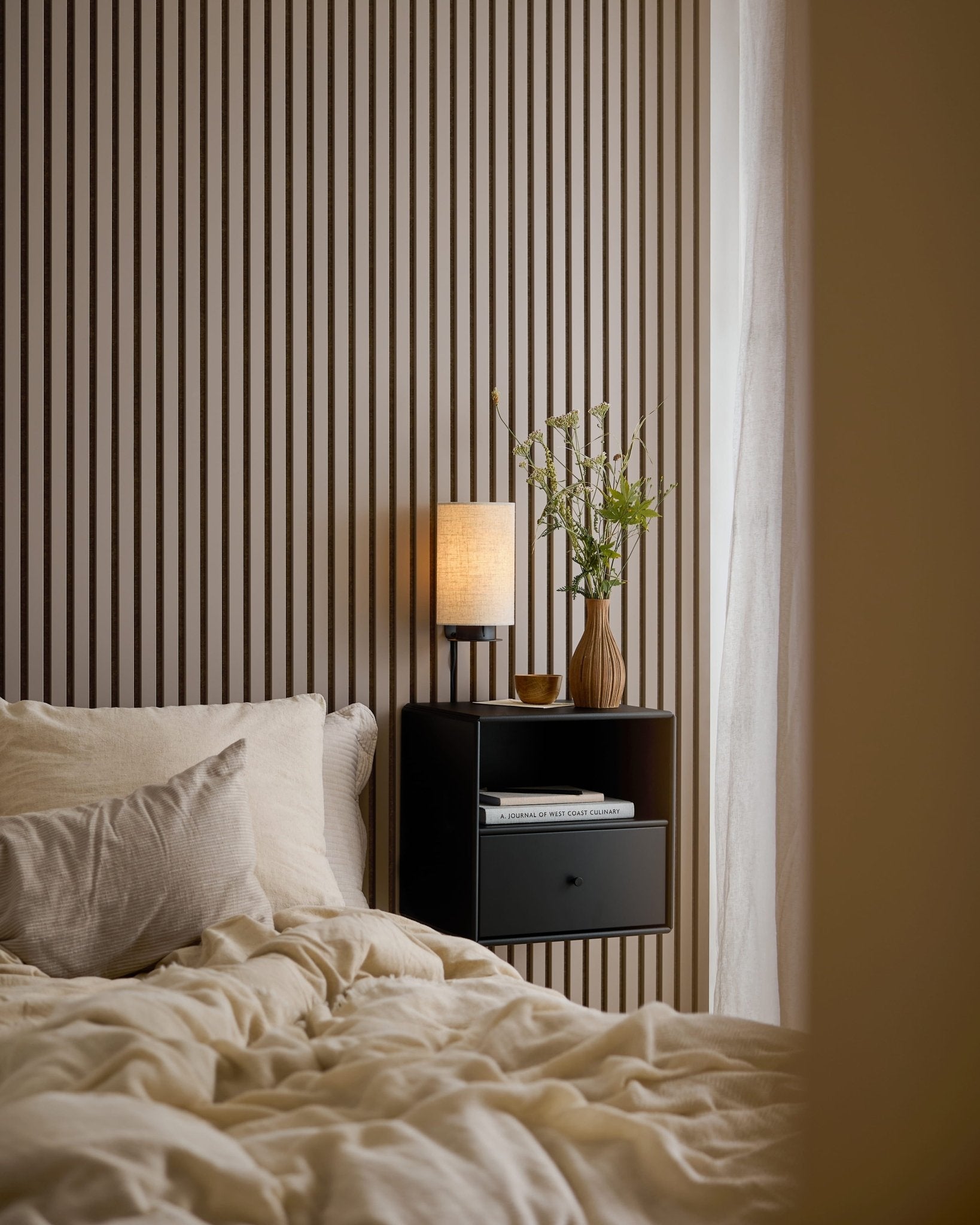 Un letto con biancheria color crema accanto a un comodino nero, davanti a una parete Akupanel in Dark Cashmere.