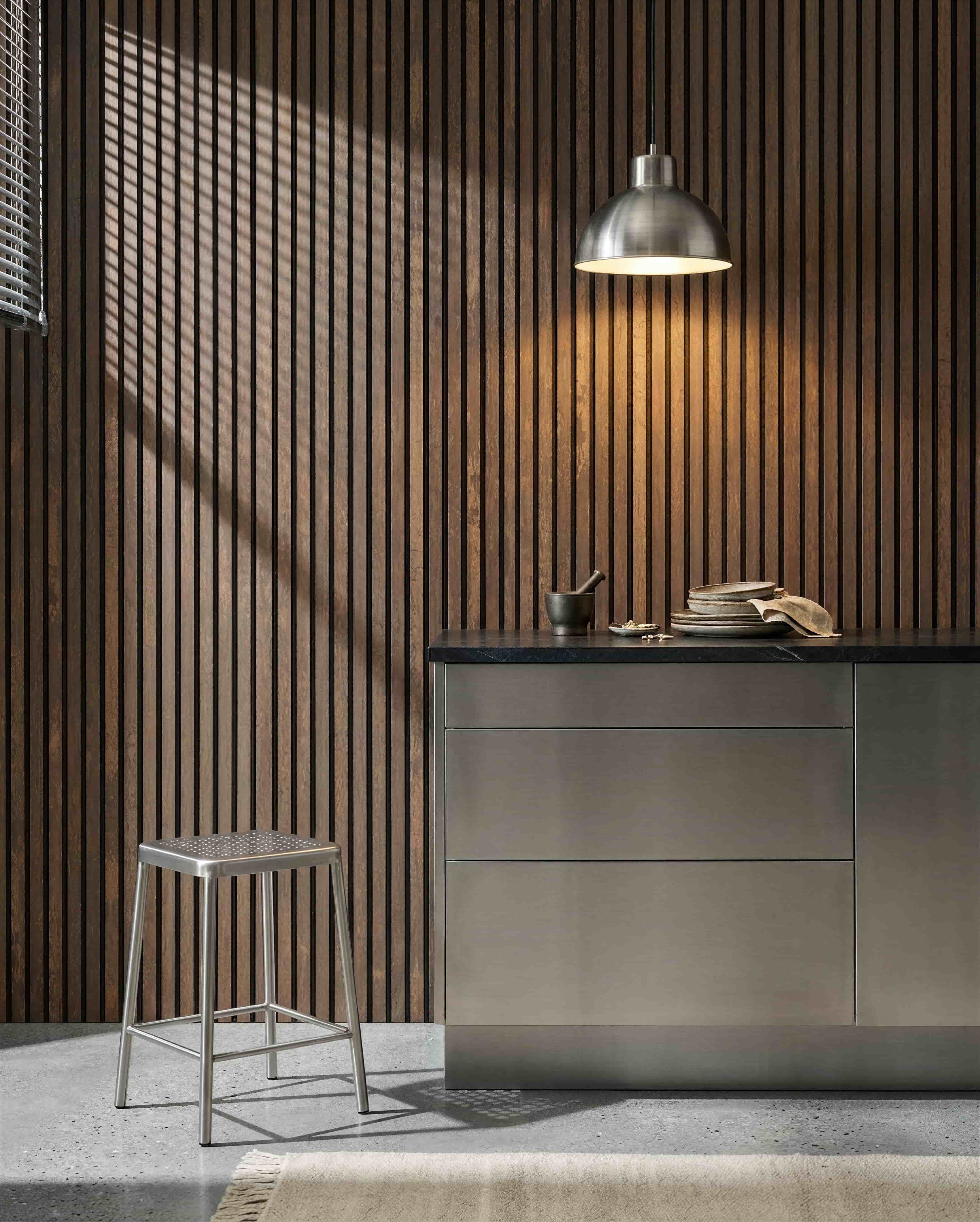 Cucina con una parete acustica a doghe WoodUpp Akupanel Oxide Copper, piano di lavoro in acciaio inox, sgabello in metallo, lampada a sospensione, piatti assortiti e mortaio con pestello.
