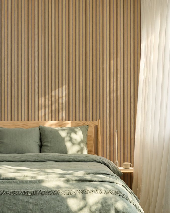 Camera da letto nordica minimalista con una parete WoodUpp Akupanel Rovere Pannello acustico, biancheria da letto verde salvia, struttura del letto in legno e tende bianche velate.
