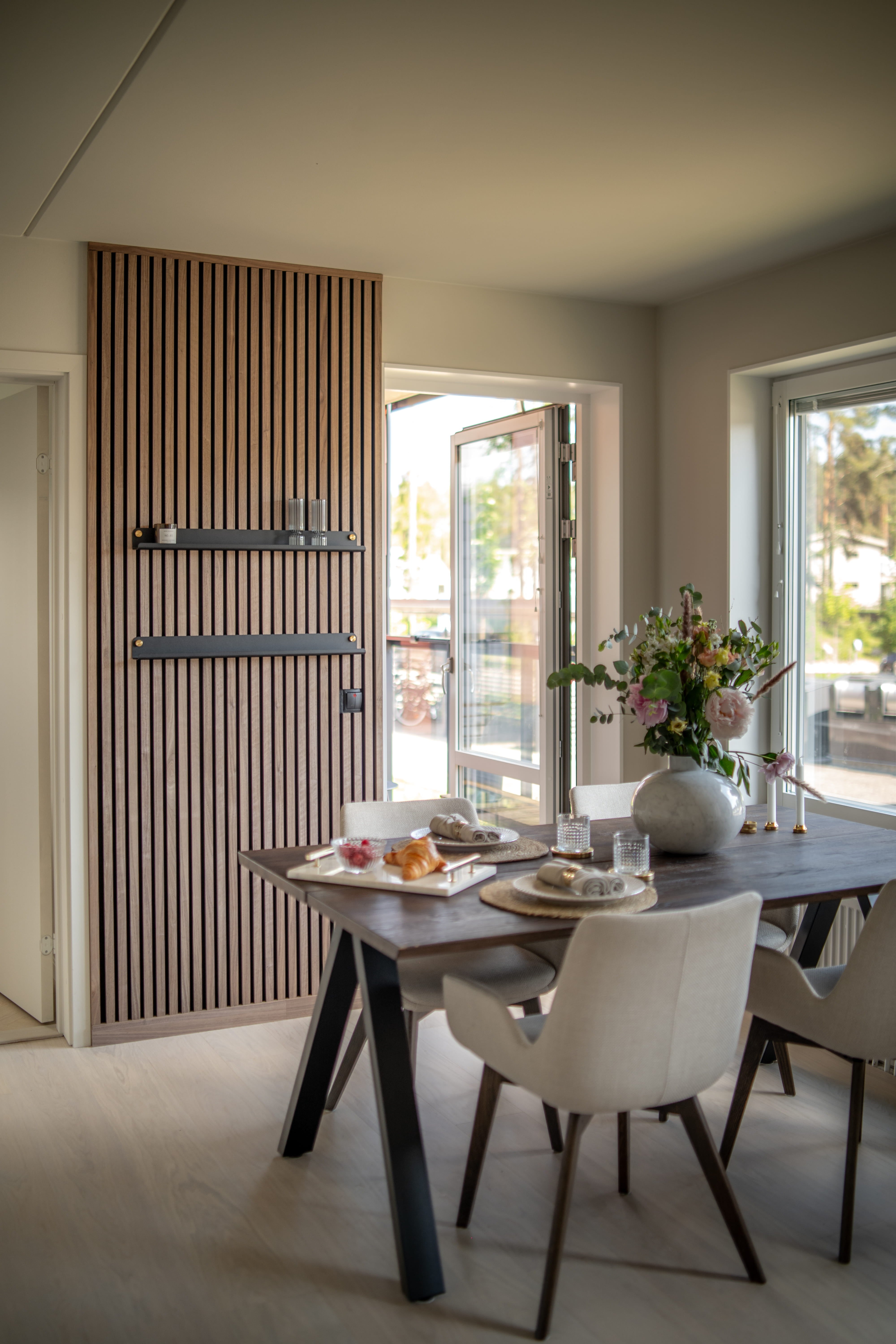 Sala da pranzo moderna con parete a doghe in legno acustico Akupanel, tavolo da colazione scuro, sedie beige minimaliste e un vaso di fiori accanto a grandi finestre aperte.