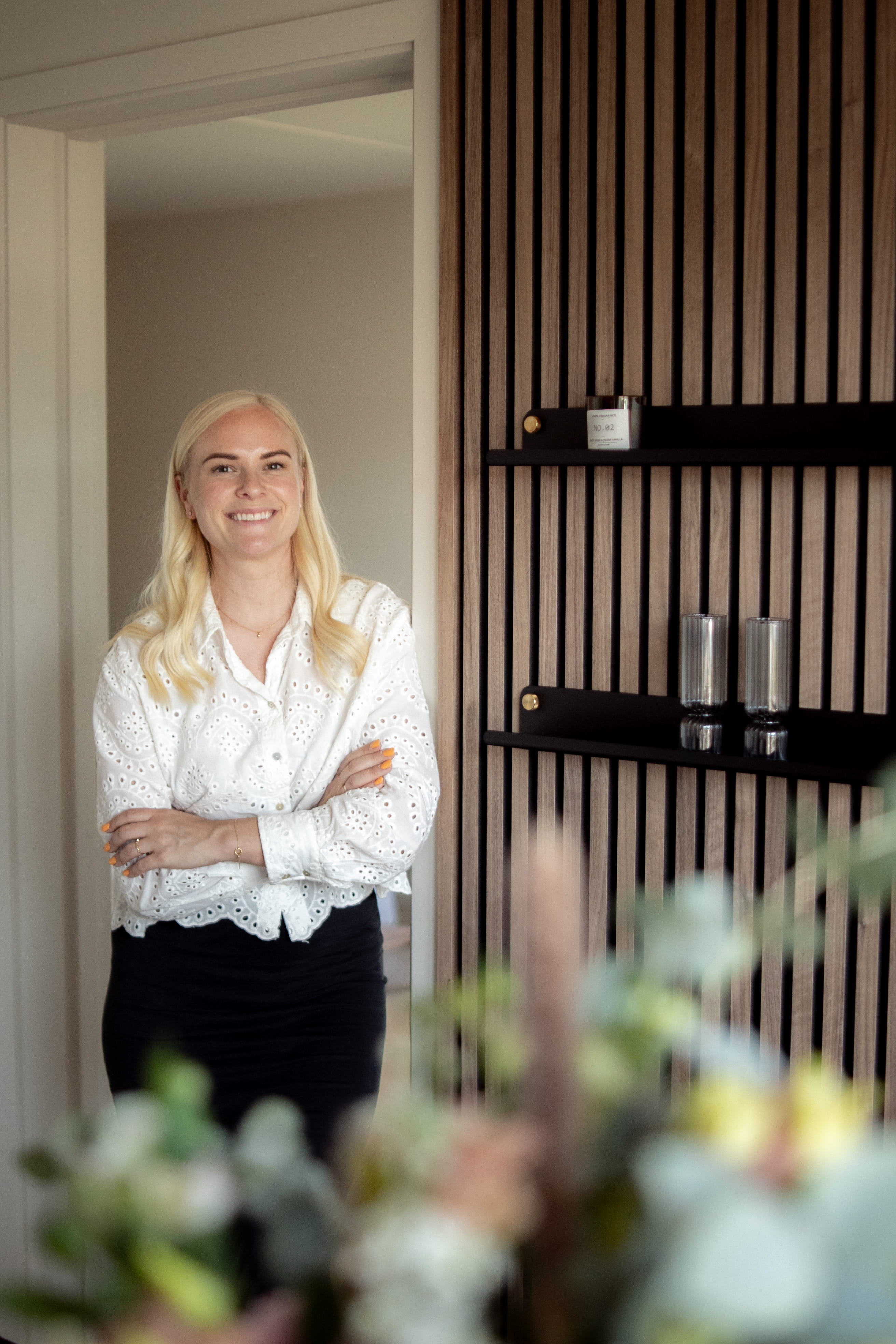 Una donna con lunghi capelli biondi e una camicetta bianca se ne sta sorridente a braccia incrociate in una stanza moderna con pannelli in legno acustico nordico e scaffali eleganti.