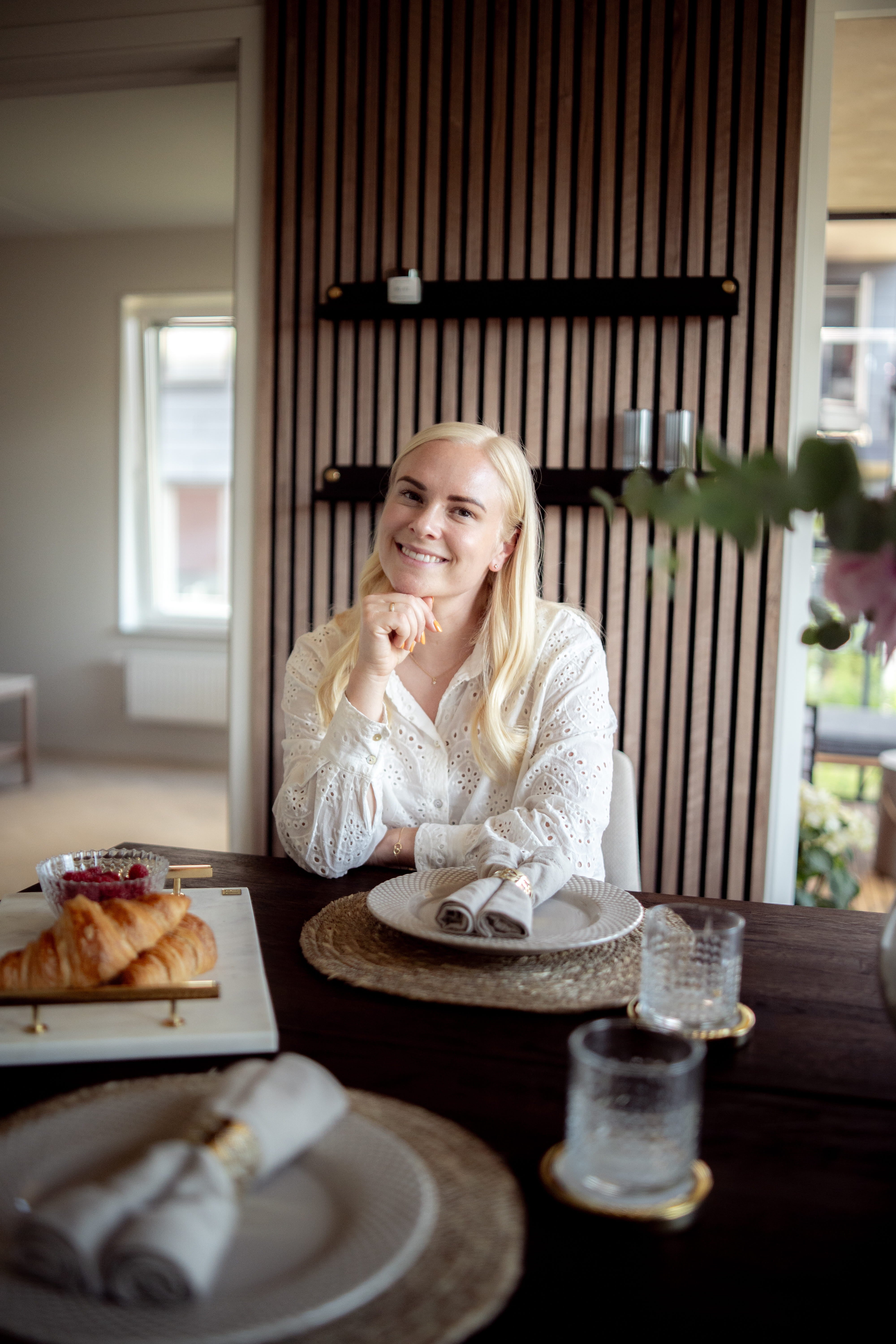 Una donna dai lunghi capelli biondi siede a un tavolo da pranzo apparecchiato per la colazione con croissant e bicchieri, accanto a un pannello acustico nordico appeso alla parete.