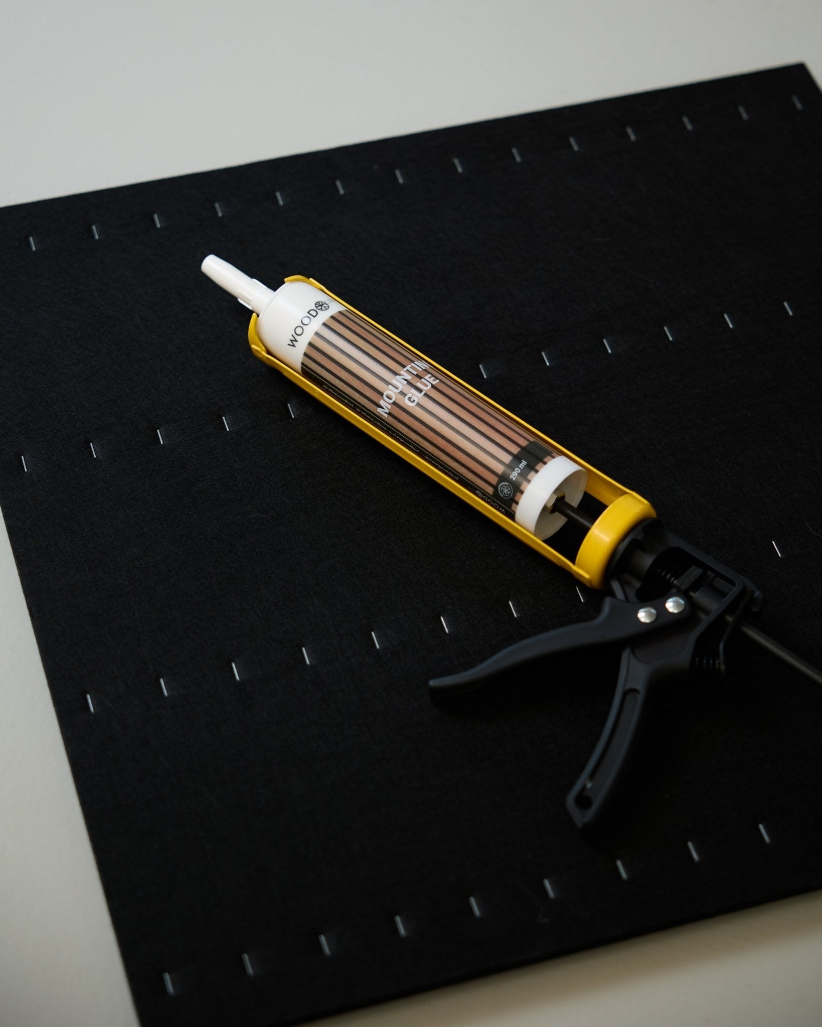 Una WoodUpp Pistola per Colla gialla e black (290 ml) è appoggiata su un tappetino black con una griglia di graffe bianche, usata per installare pannelli acustici.