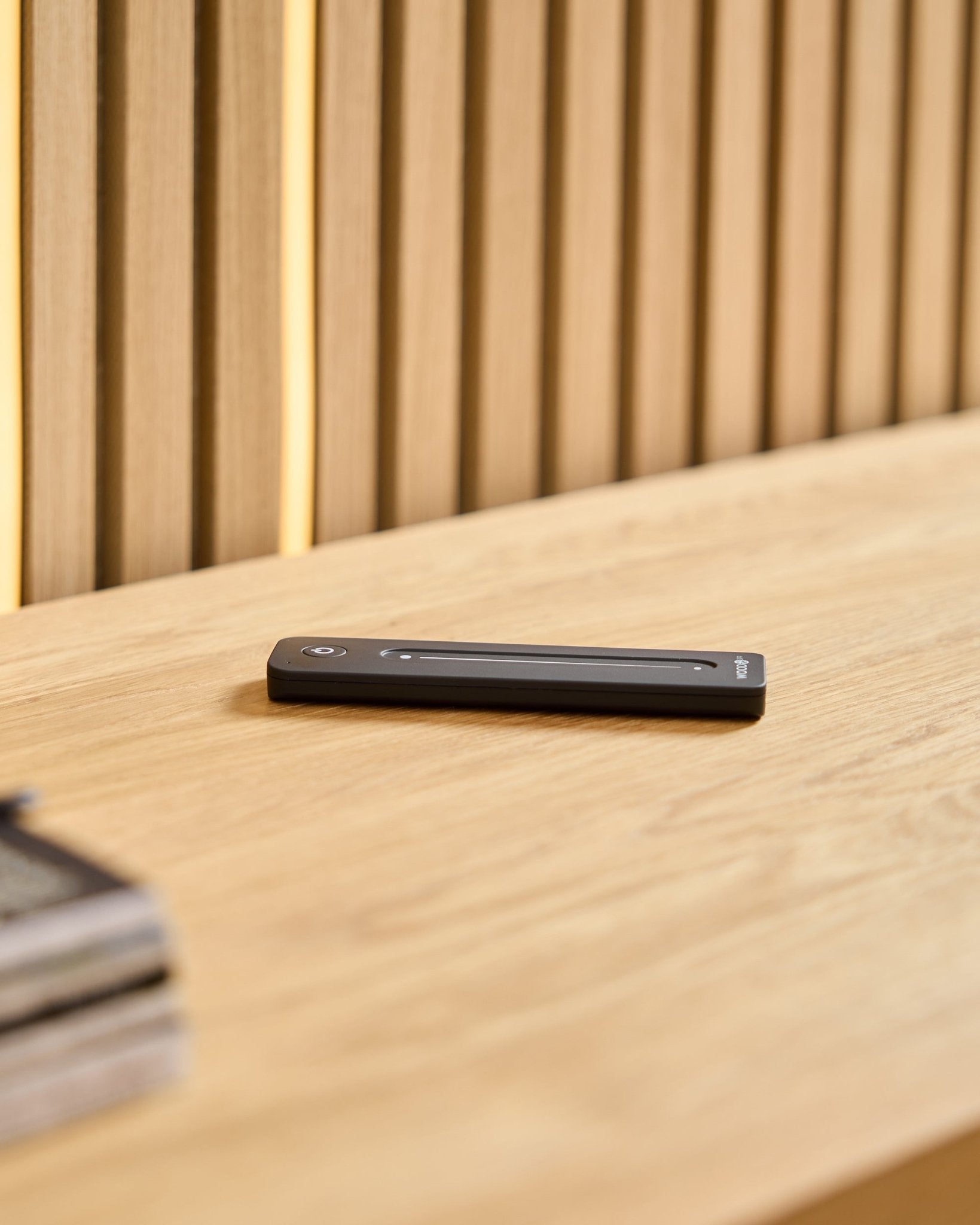 Un telecomando LED black WoodUpp Lumi su una superficie in legno chiaro, con sullo sfondo pannelli acustici in legno a doghe.