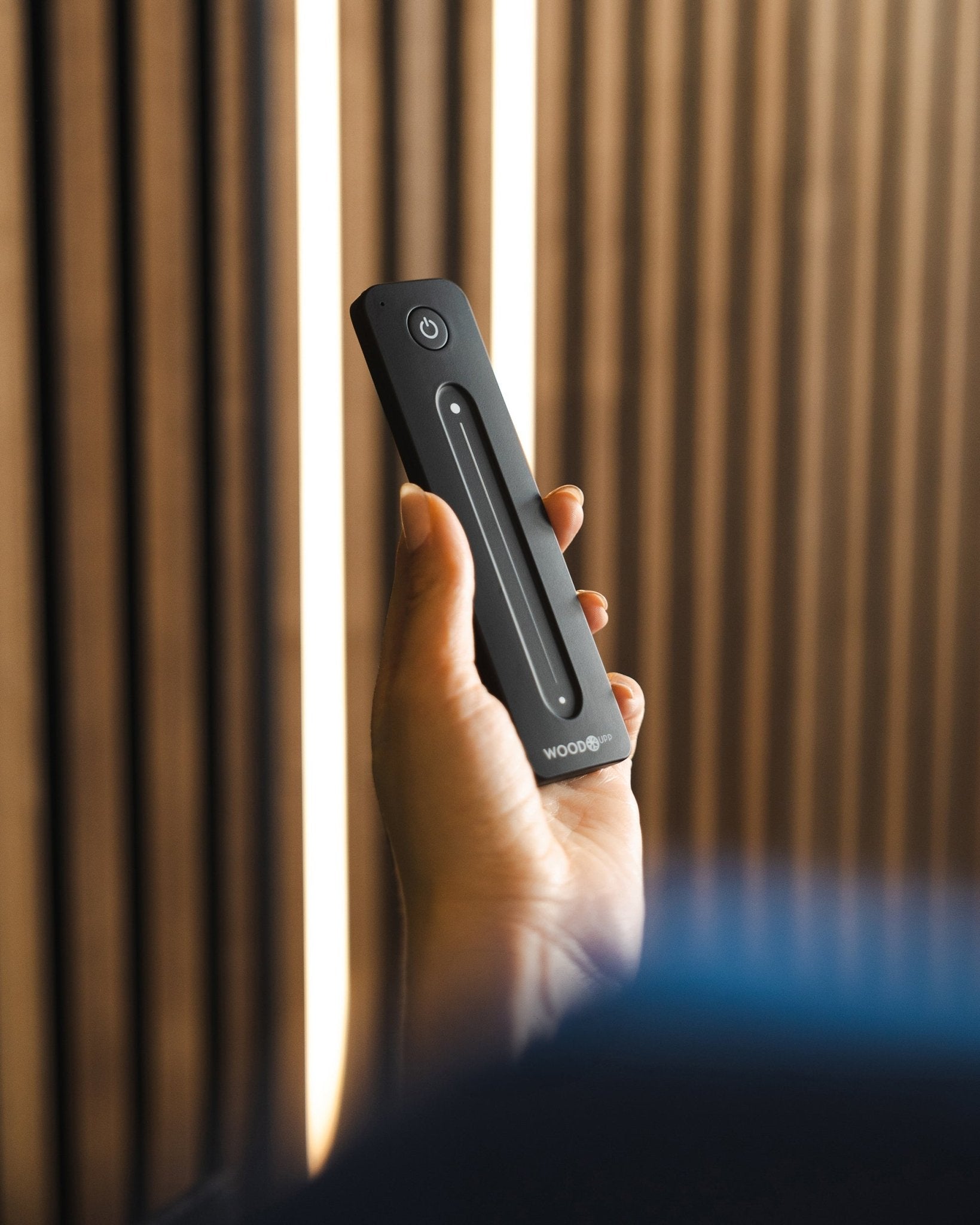 Una mano tiene in mano un telecomando per strip LED black WoodUpp Lumi con pulsante di accensione, posizionato davanti ai pannelli acustici a doghe WoodUpp; compatibile con Philips Hue.