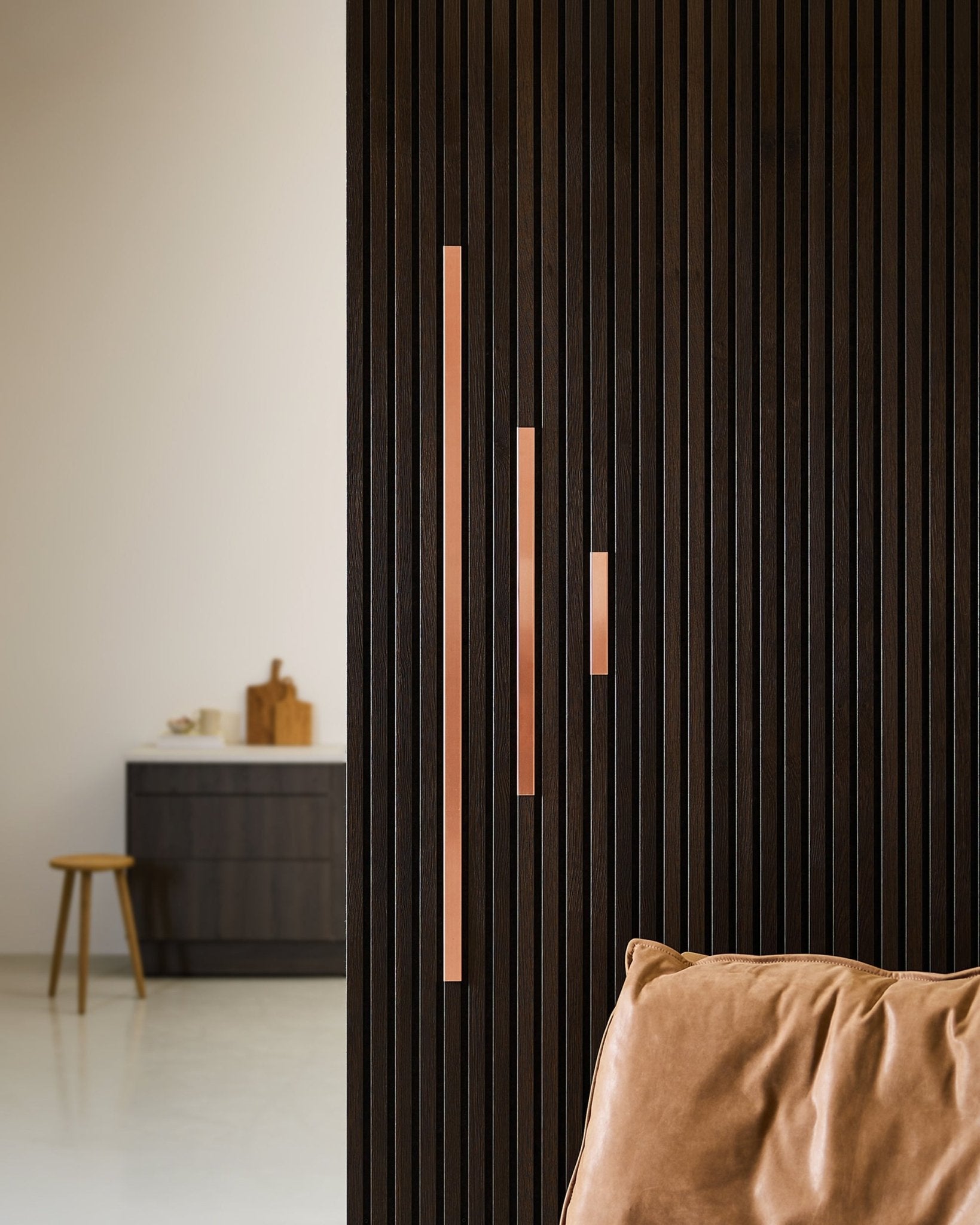 Spazio minimalista con una parete divisoria acustica a doghe in legno scuro, con pannelli WoodUpp Liva Cover in copper, 60 cm con maniglie copper, poltrona in pelle color cuoio e sgabello contenitore.
