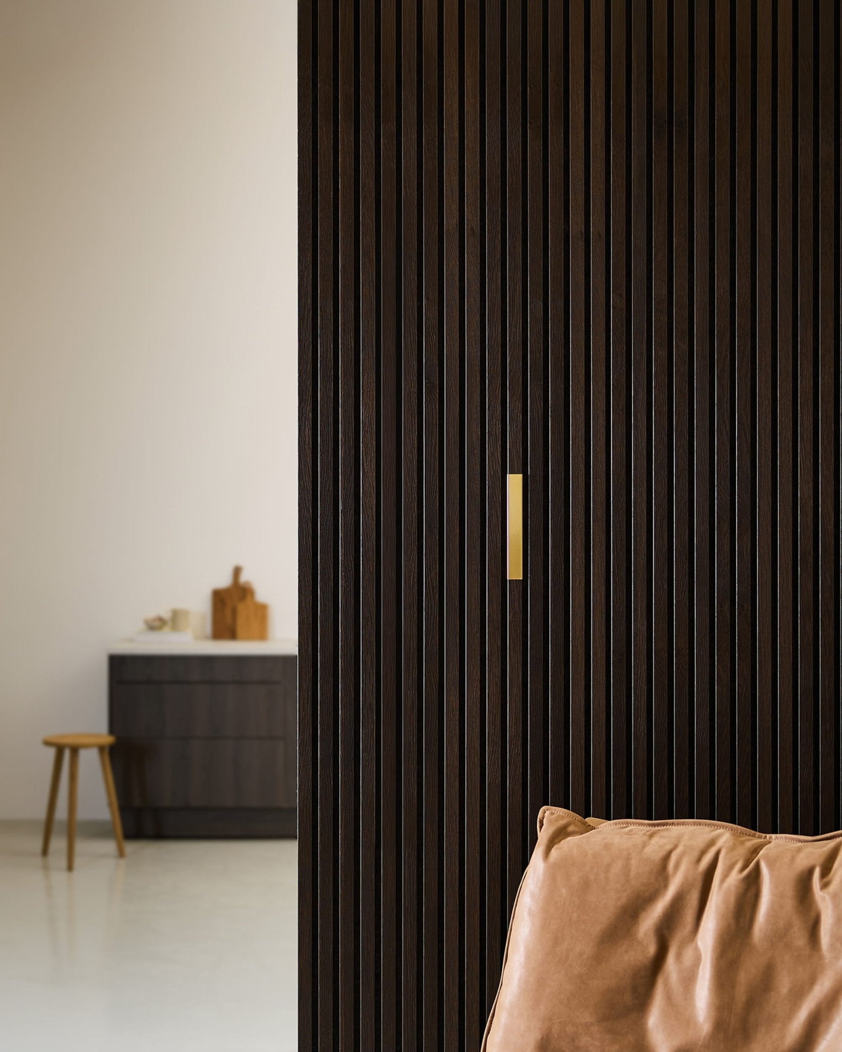 Stanza in stile minimalista con un elemento WoodUpp Liva | Cover / Brass da 20 cm su una parete divisoria Akupanel in legno scuro, una poltrona in pelle marrone e un mobile nero.
