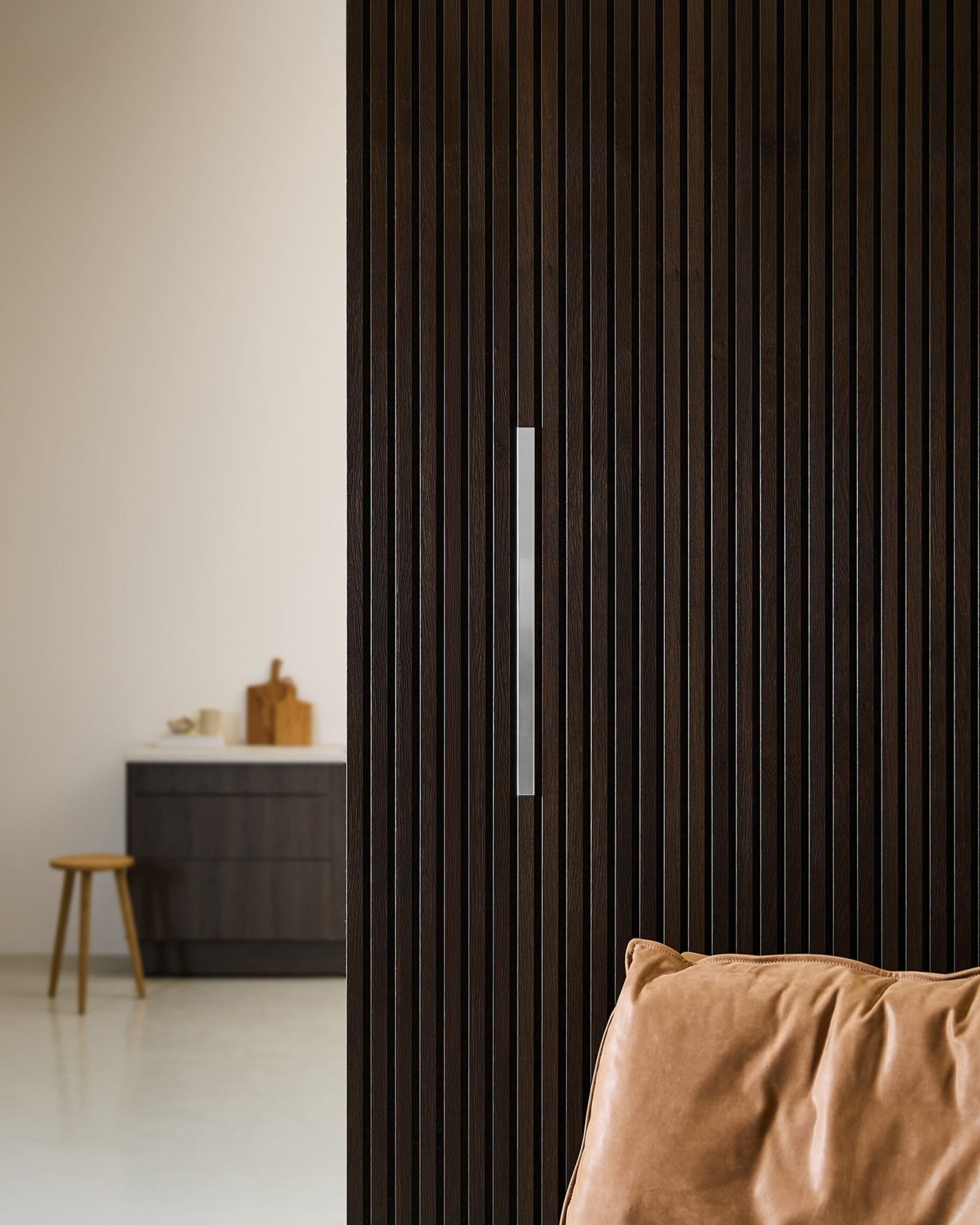 Spazio in stile nordico moderno con pannelli acustici WoodUpp Liva in acciaio lucido, una parete a doghe in legno scuro, una poltrona in pelle color cuoio e uno sgabello accanto a un mobile da cucina.