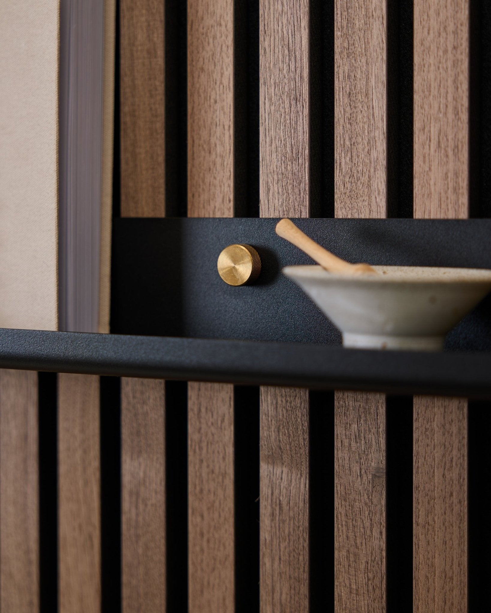 Primo piano di una mensola stretta WoodUpp Norr black da 115,9 cm montata su un pannello acustico a doghe nordico con viti zigrinate brass, pomello dorato e ciotola in ceramica con cucchiaio.
