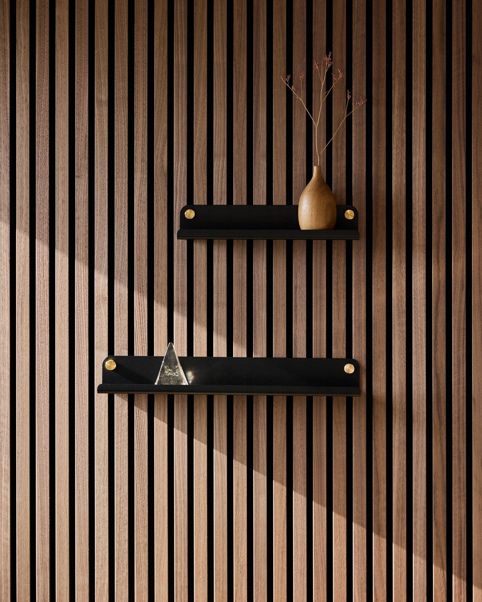 Due mensole strette WoodUpp Norr black (35,9 cm) montate su un pannello acustico a doghe in stile nordico; sulla mensola superiore c’è un vaso con rami secchi, su quella inferiore una piramide di vetro.