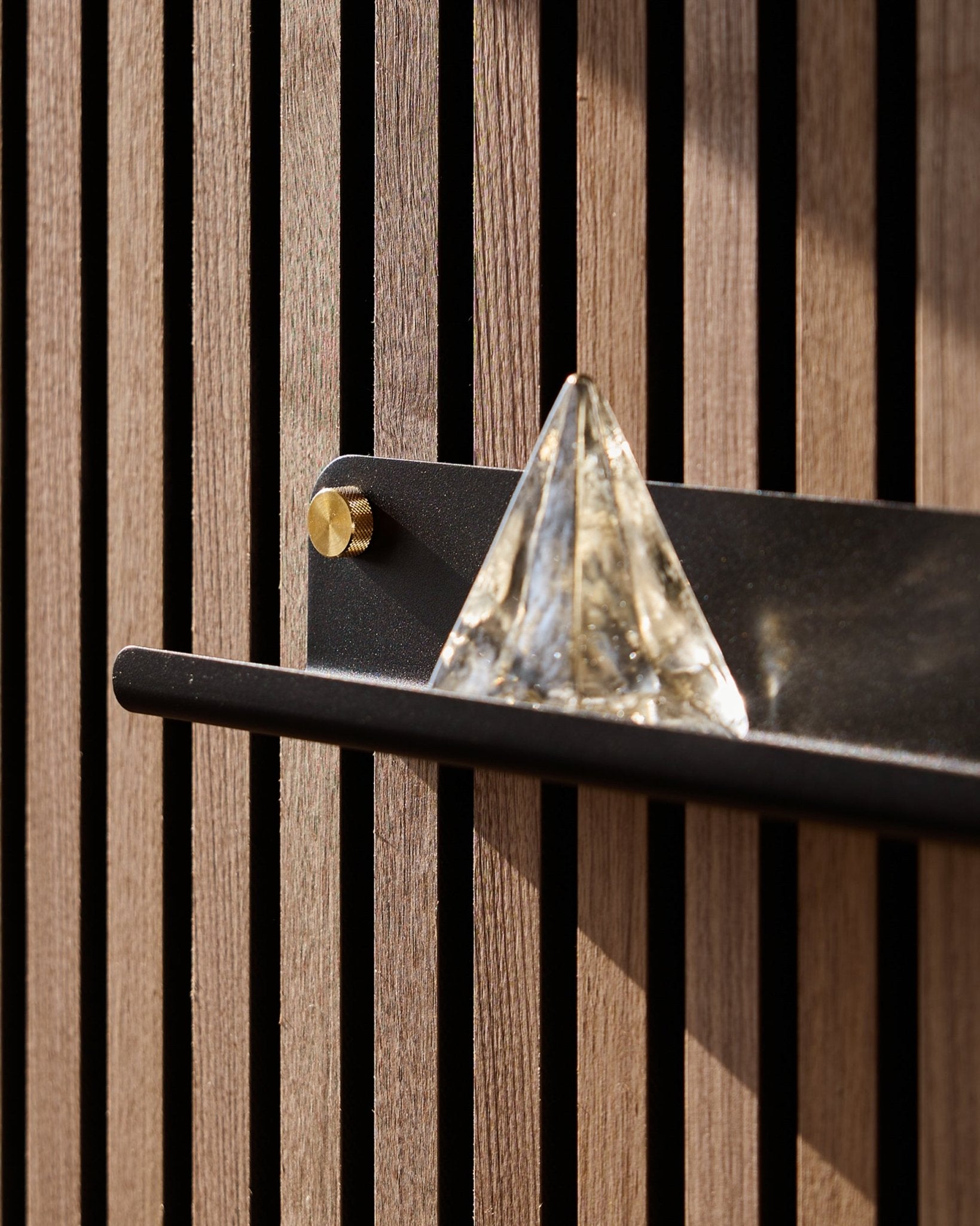 Una piramide in vetro è appoggiata sulla WoodUpp Norr Narrow Shelf in black (51,9 cm), montata su una vertical Pannello acustico e valorizzata dalla luce del sole.