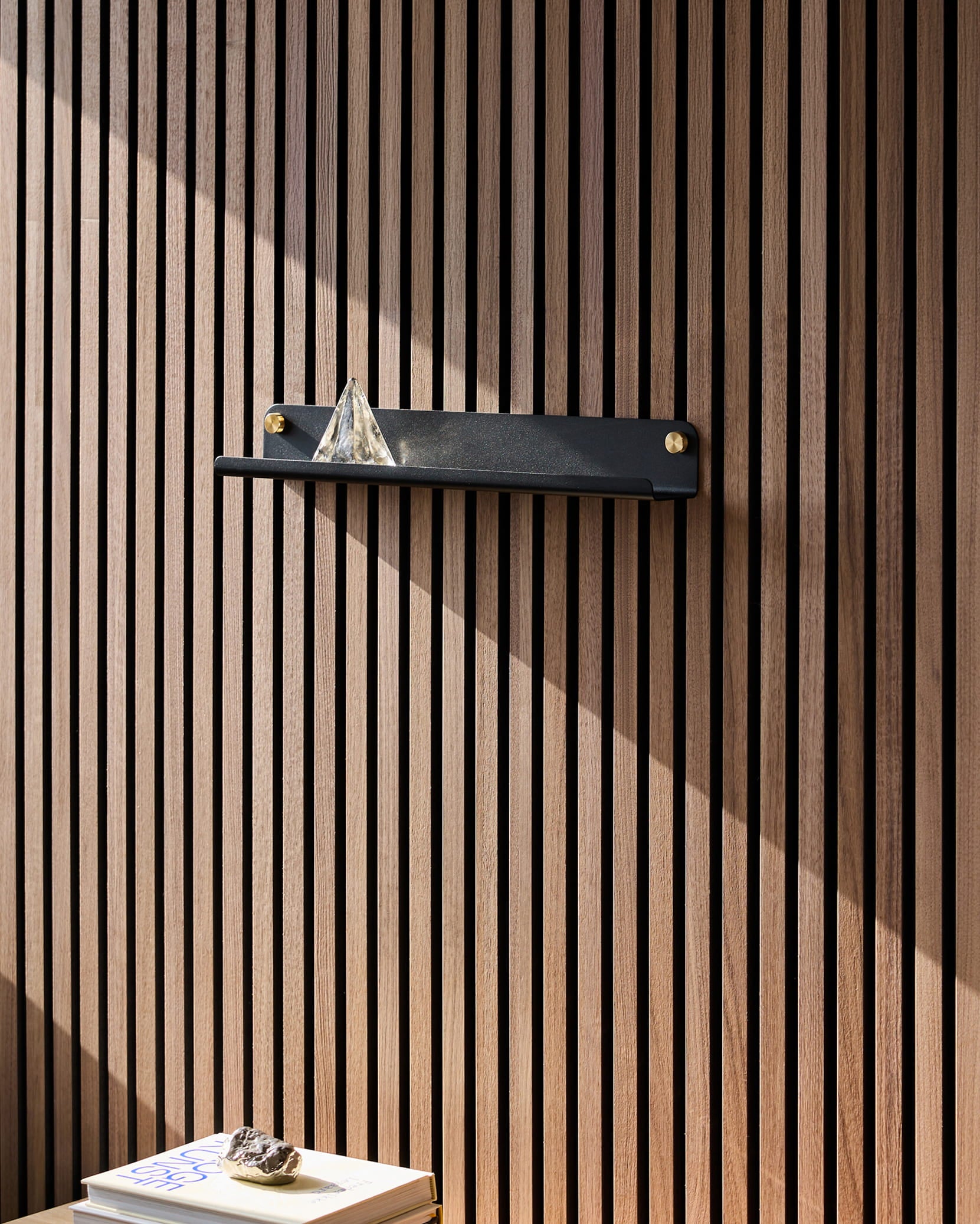 Una mensola Norr Narrow Shelf nera di WoodUpp, appoggiata su un pannello acustico in legno Akupanel, sostiene una piramide di vetro; la luce del sole illumina i libri sottostanti, su cui è posata una pietra.