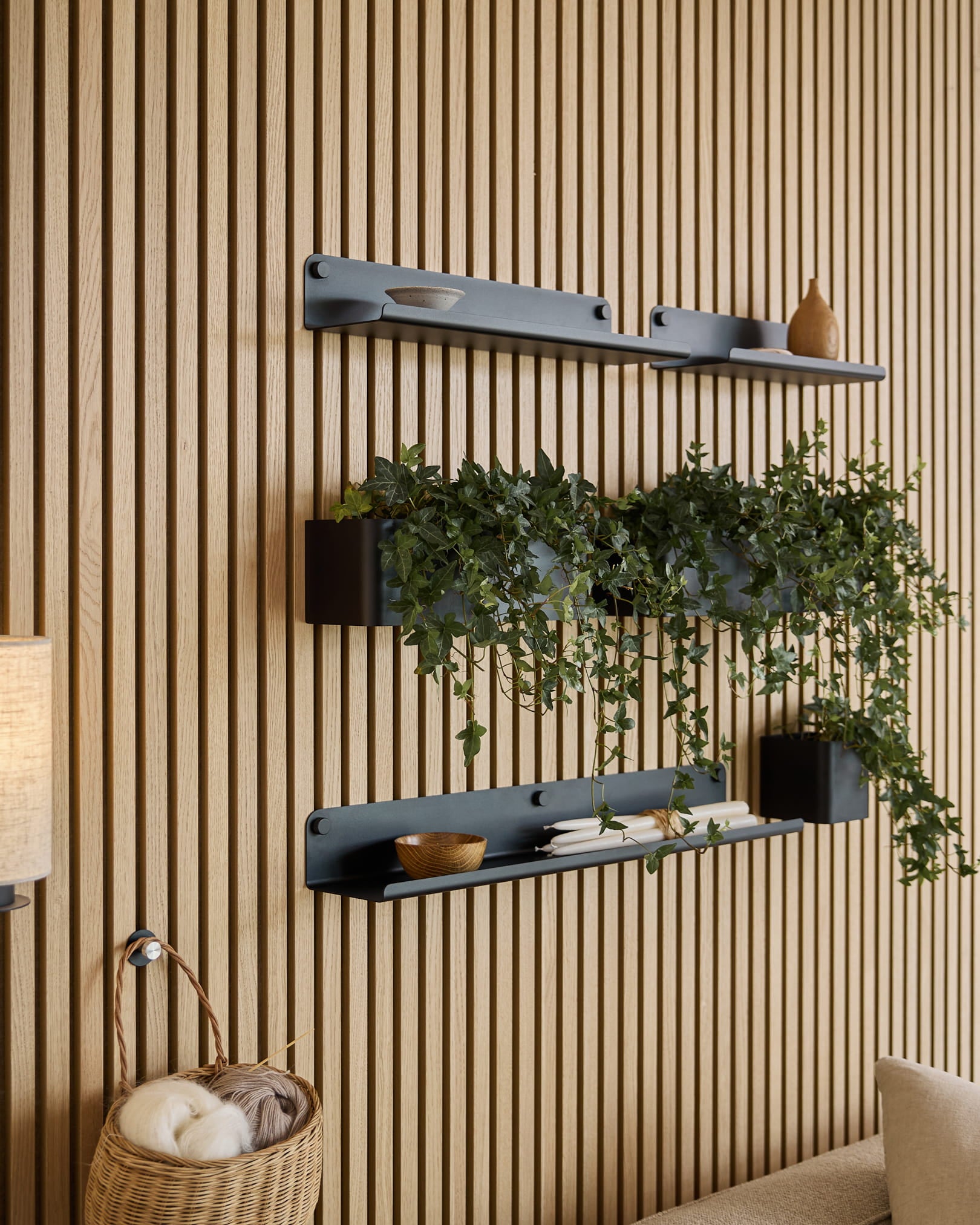 Tre mensole WoodUpp black Norr | Plant Box con oggetti decorativi e una pianta sono montate sopra un cesto in vimini con gomitoli, su un pannello fonoassorbente a doghe verde.