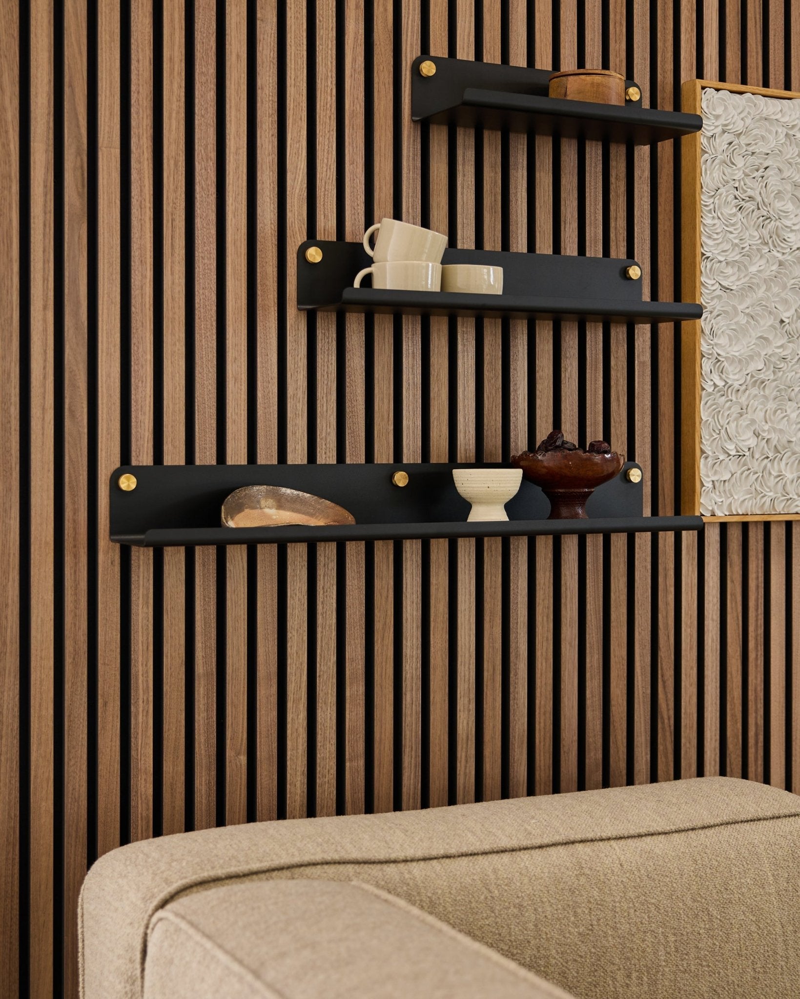 Tre mensole black, tra cui la mensola larga WoodUpp Norr (75,9 cm), espongono oggetti decorativi su una parete Pannello acustico in doghe di legno sopra un divano beige; opera d’arte parzialmente visibile.