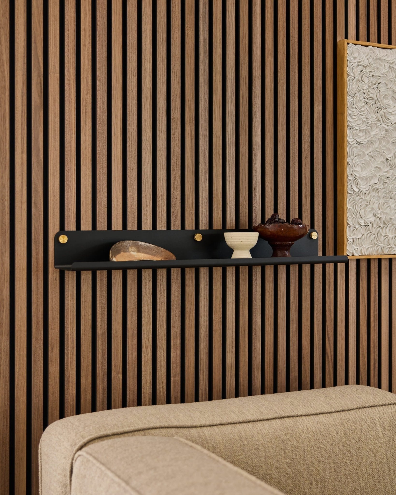 Mensola larga Black WoodUpp Norr (75,9 cm) con decorazione sopra un divano beige, fissata a un moderno pannello acustico in legno a doghe.