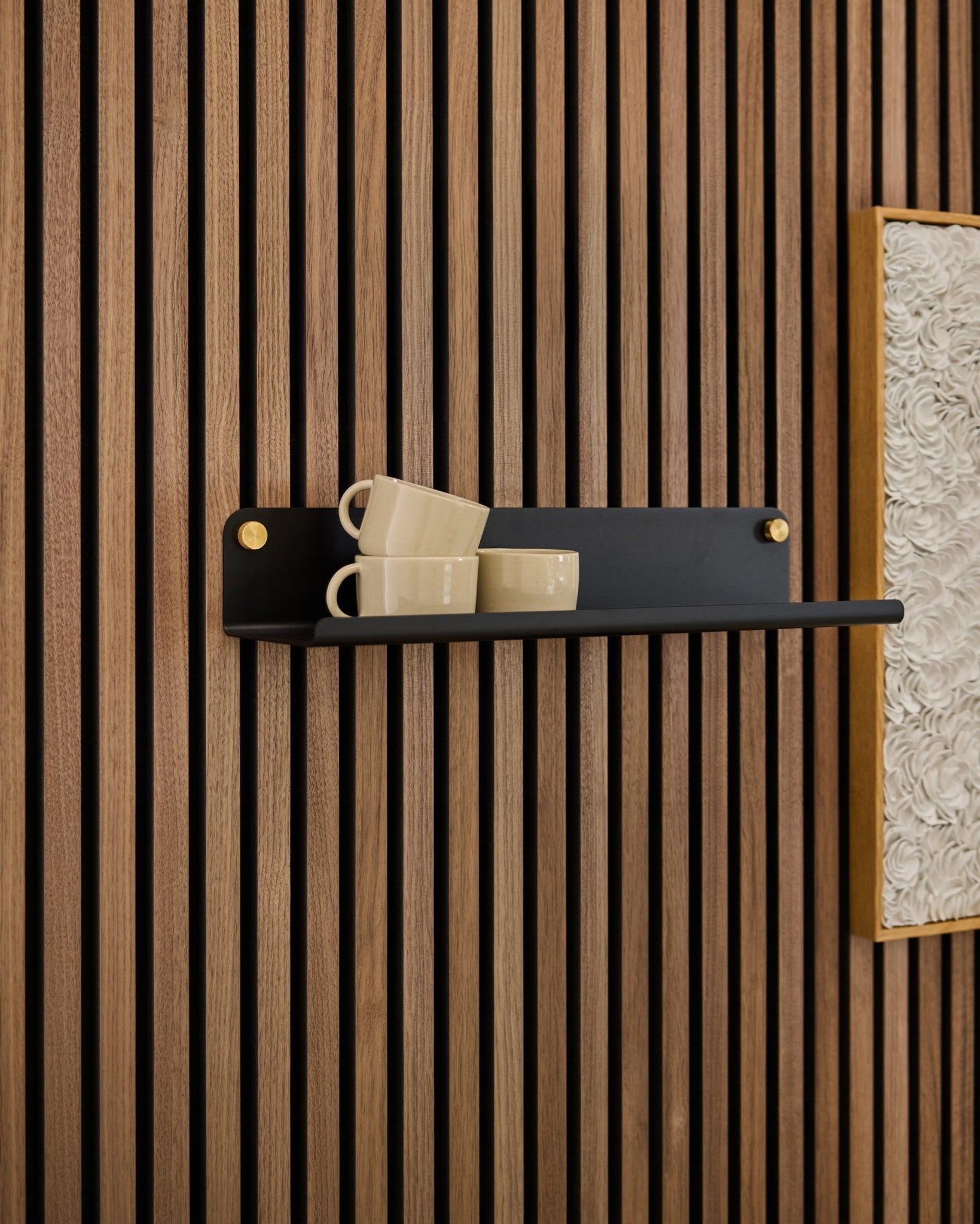 Una mensola Norr Wide Shelf nera di WoodUpp ospita tre tazze beige, fissate su un pannello acustico Akupanel accanto a un’opera d’arte bianca incorniciata dalla superficie ruvida.