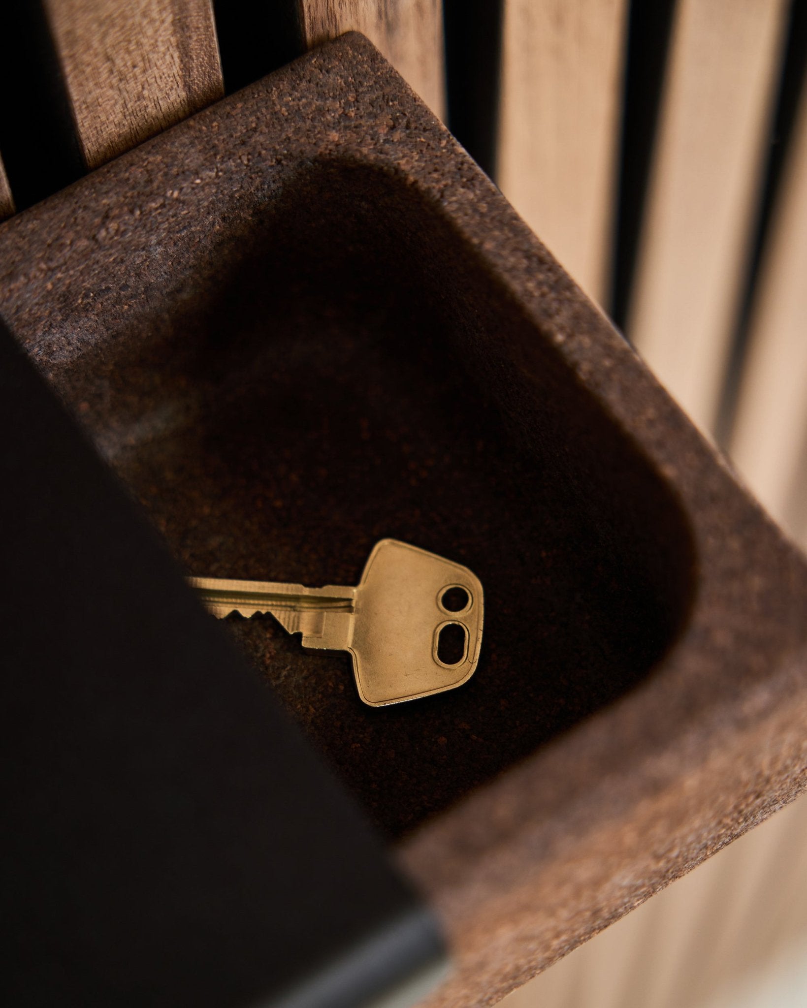 Una chiave brass è appoggiata su un Norr Key Tray/termotrattato Cork, 35,9 cm, di WoodUpp, posizionato su un contenitore brown davanti a uno sfondo di pannelli acustici in legno Akupanel.