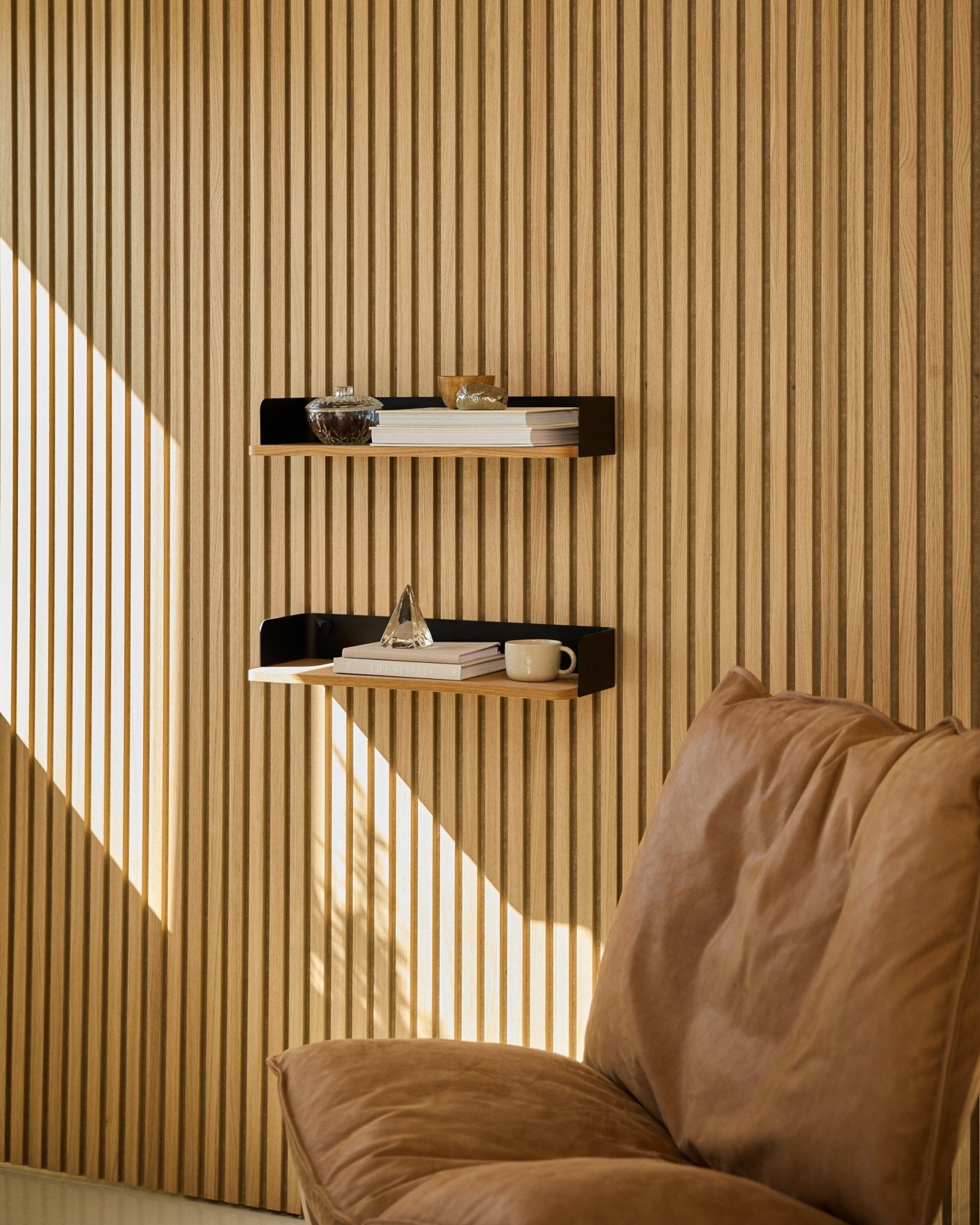 Sedia Brown accanto alla libreria WoodUpp Linn in Natural Oak, con libri e oggetti decorativi disposti nella luce naturale davanti a una parete a listelli in legno nordico Pannello acustico.