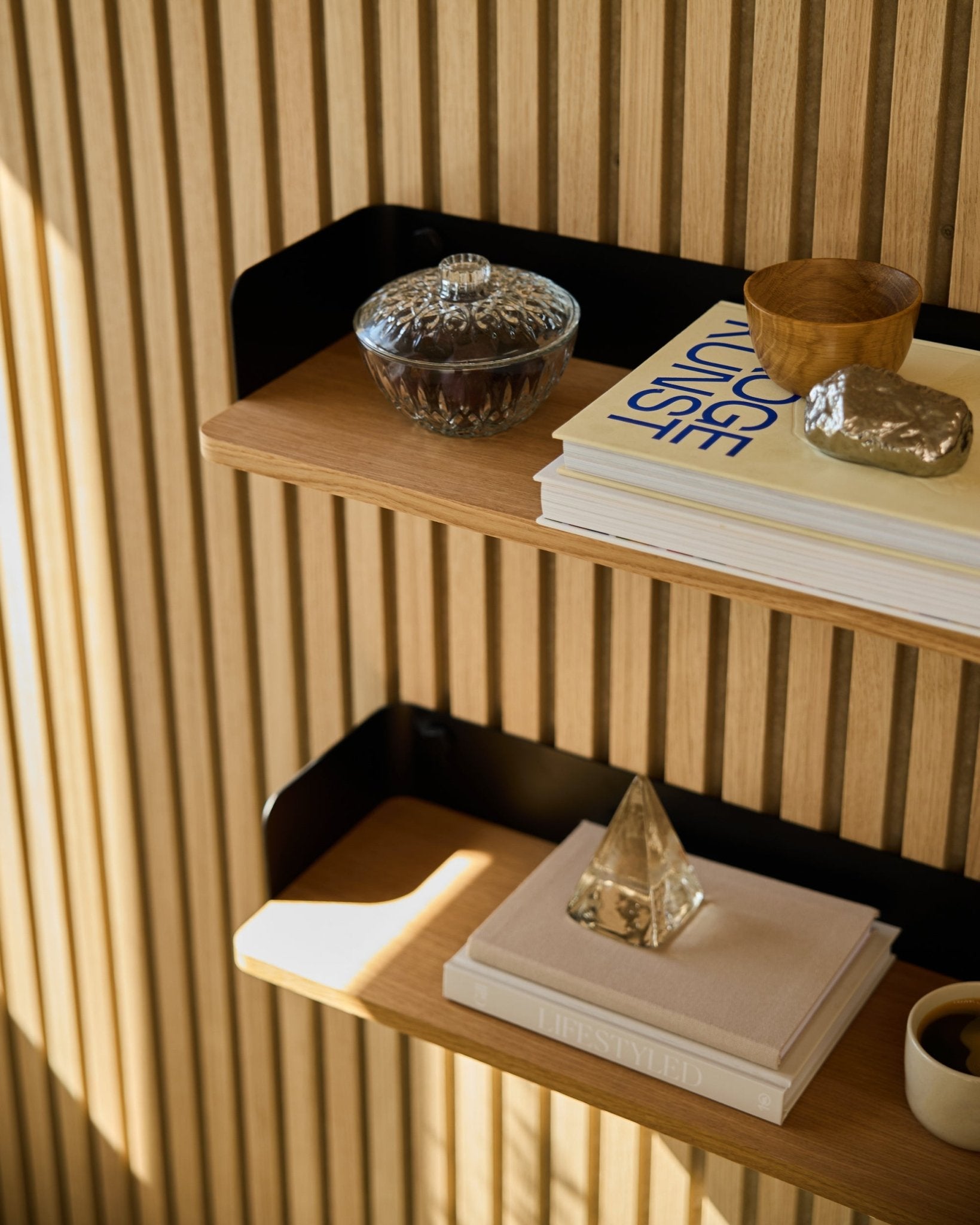 La libreria WoodUpp Linn in Natural Oak (55,9 cm) ospita un barattolo in vetro, una tazza, una piramide dorata e libri davanti a pannelli acustici in legno Akupanel illuminati dal sole.