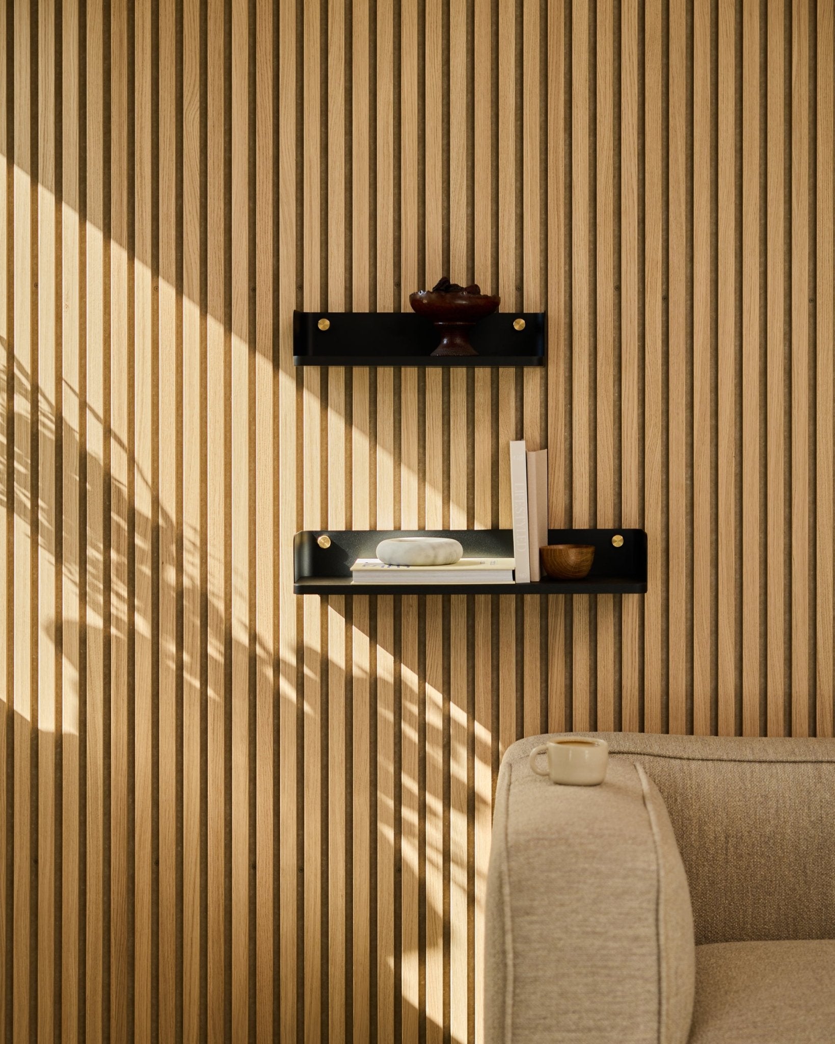 La luce del sole illumina un divano beige e la libreria Linn di WoodUpp in black oak, 55,9 cm, su uno sfondo minimalista di pannello a doghe nordico Pannello acustico.