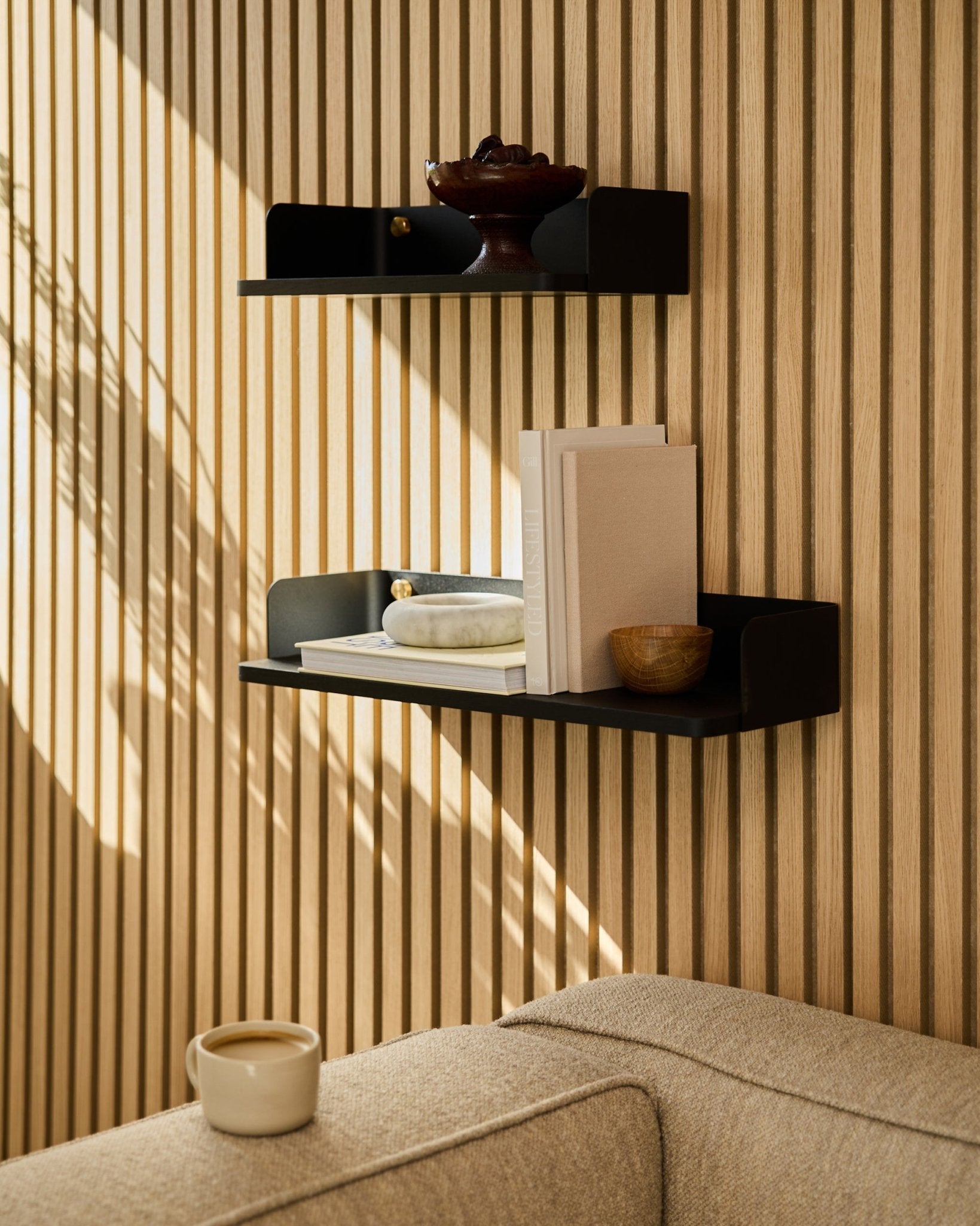 Una libreria WoodUpp black oak (55,9 cm) espone libri, oggetti decorativi e una ciotola su una parete con pannelli acustici in legno; in primo piano, un divano beige e una tazza.
