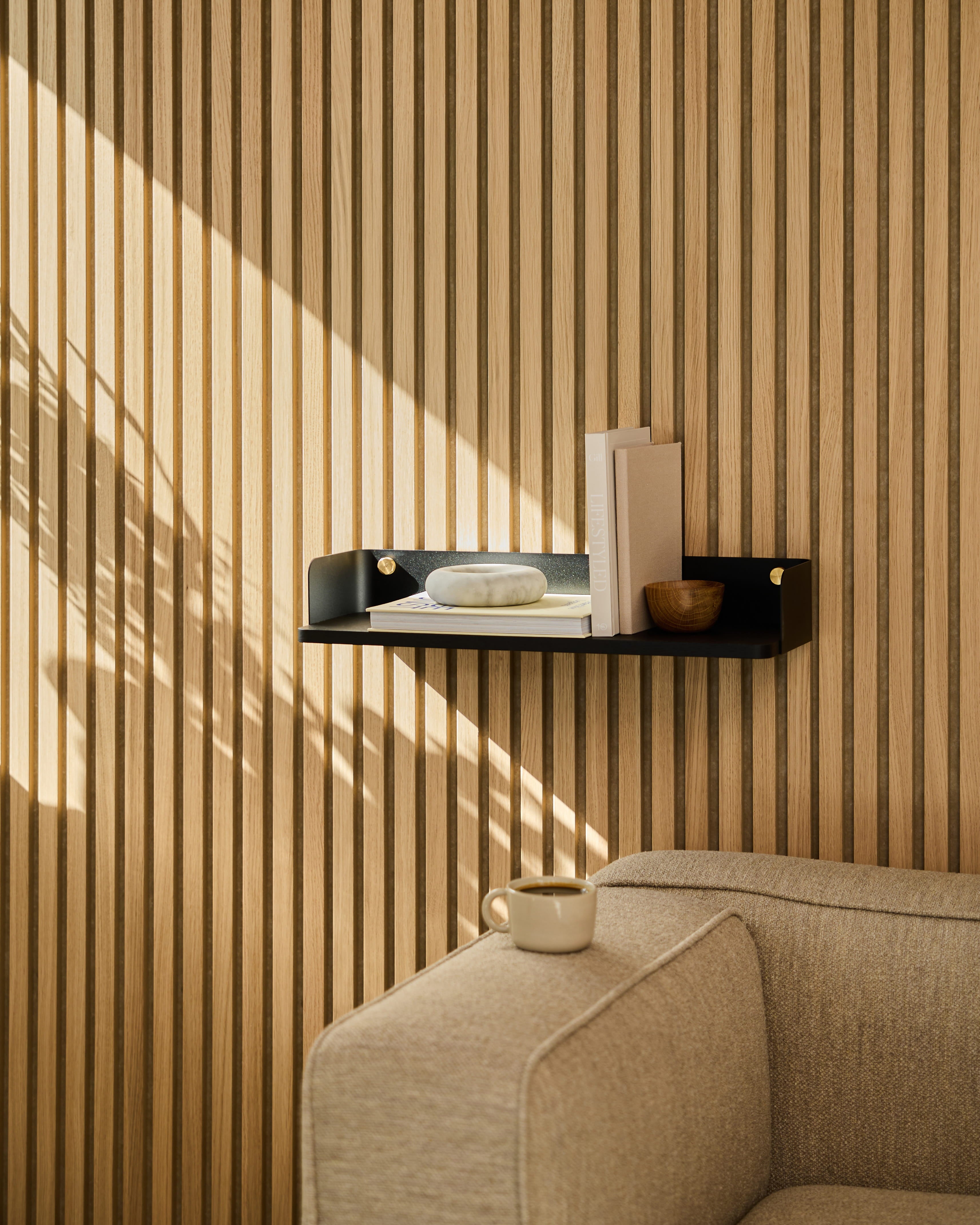 Una libreria Linn | Bookshelf / Black Oak di WoodUpp fa da cornice a un divano beige. La luce del sole proietta ombre sulla parete rivestita da pannelli acustici in legno a strisce.