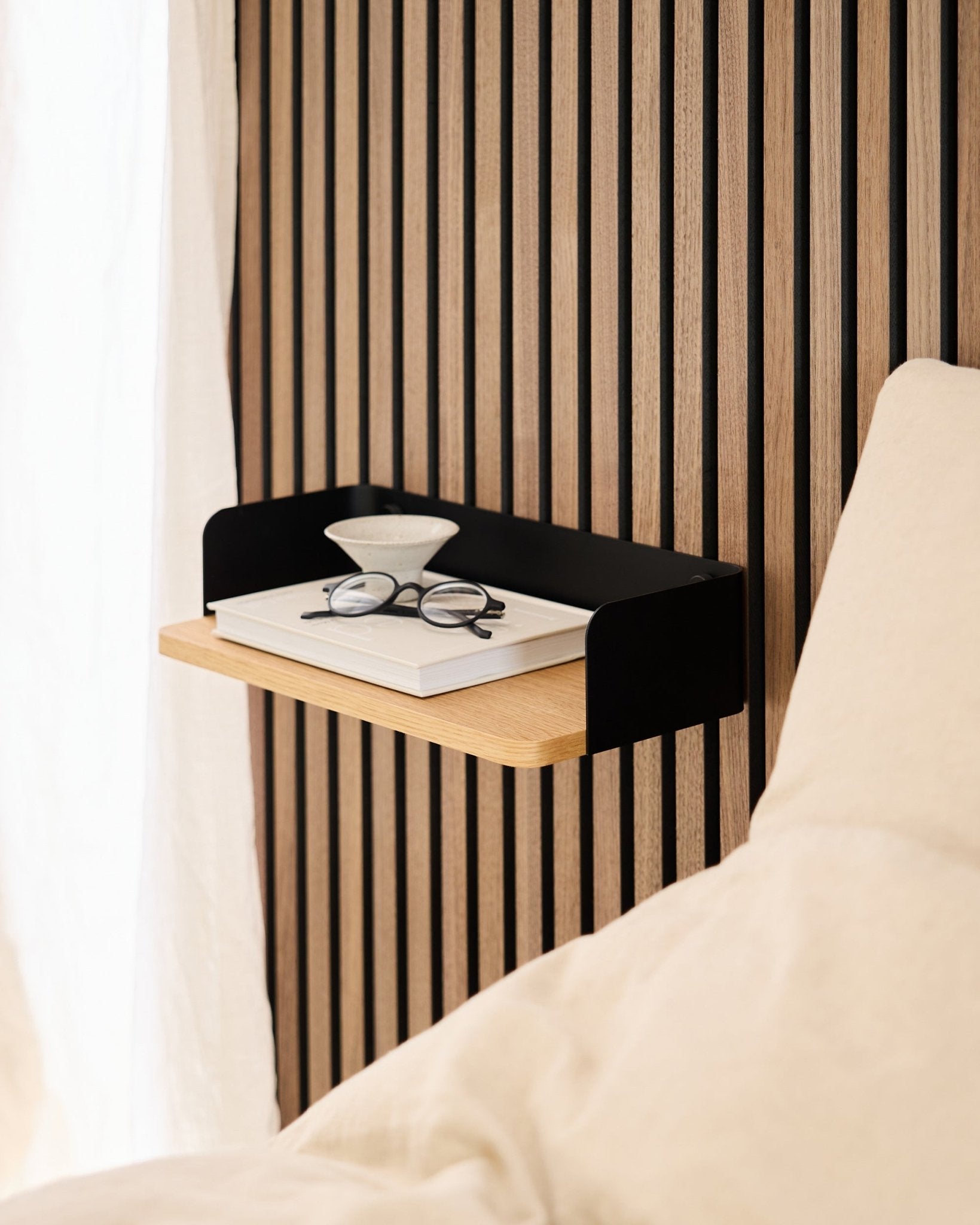 La libreria Linn di WoodUpp in natural oak (39,9 cm), con un libro, degli occhiali e un bicchiere accanto al letto, davanti a una parete con pannelli acustici in legno Akupanel.