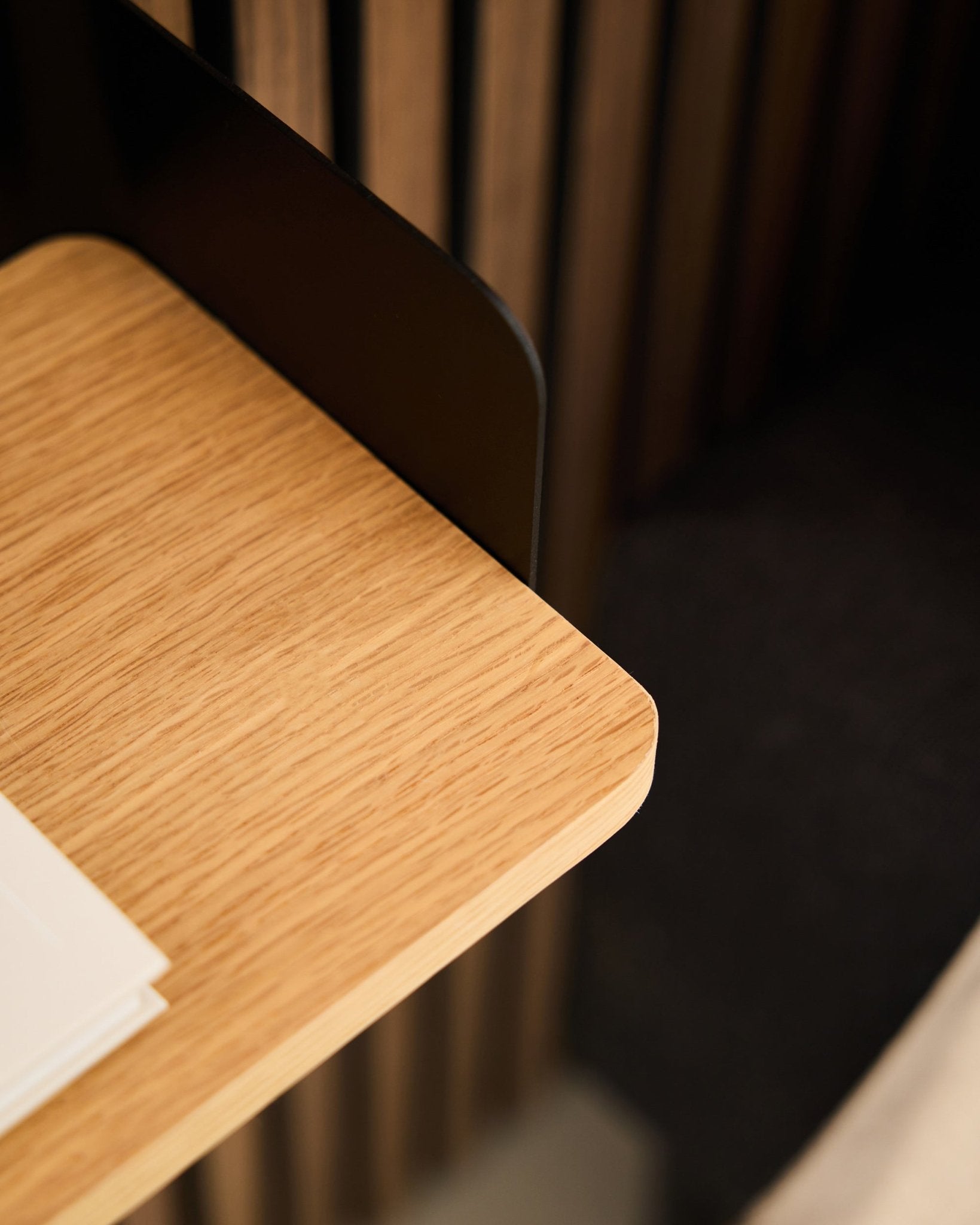 Primo piano di un angolo della scrivania con pannelli acustici in legno black Akupanel, fogli bianchi e la libreria WoodUpp Linn in Natural Oak (39,9 cm) sullo sfondo.