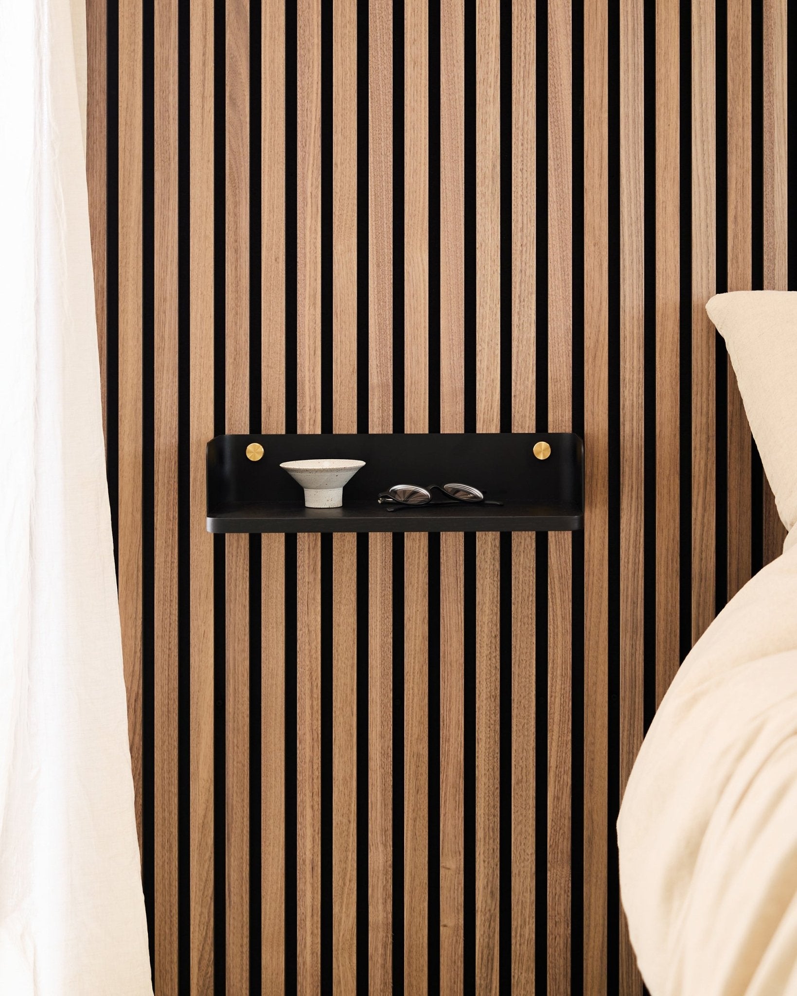 La libreria Linn in Black Oak di WoodUpp è montata su un pannello acustico a doghe in stile nordico e ospita una ciotola e dei bicchieri accanto a un letto con biancheria da letto beige.