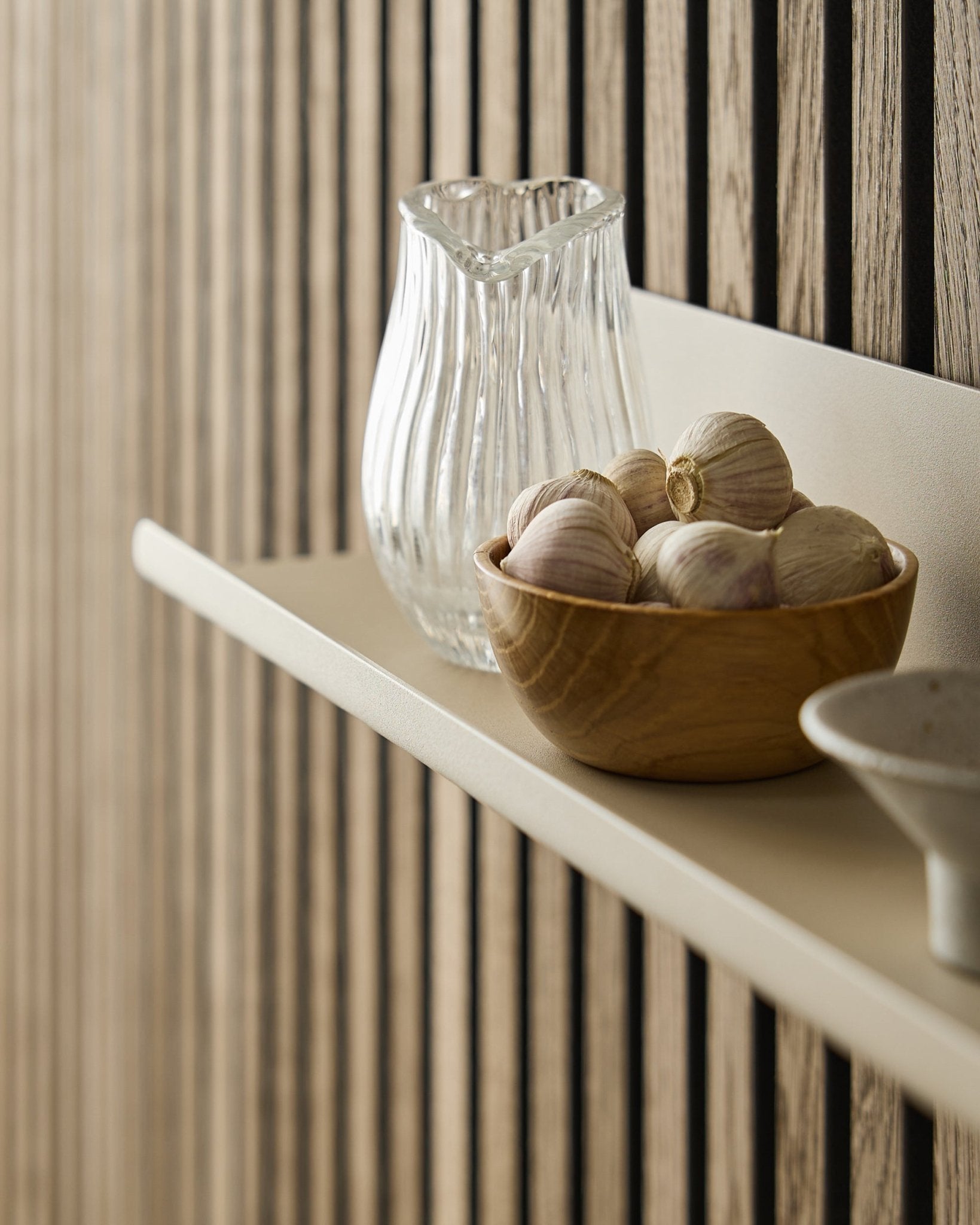 Una ciotola di legno con aglio e una brocca di vetro sulla Norr Wide Shelf di WoodUpp, montata su un Akupanel Pannello acustico con doghe in legno vertical.