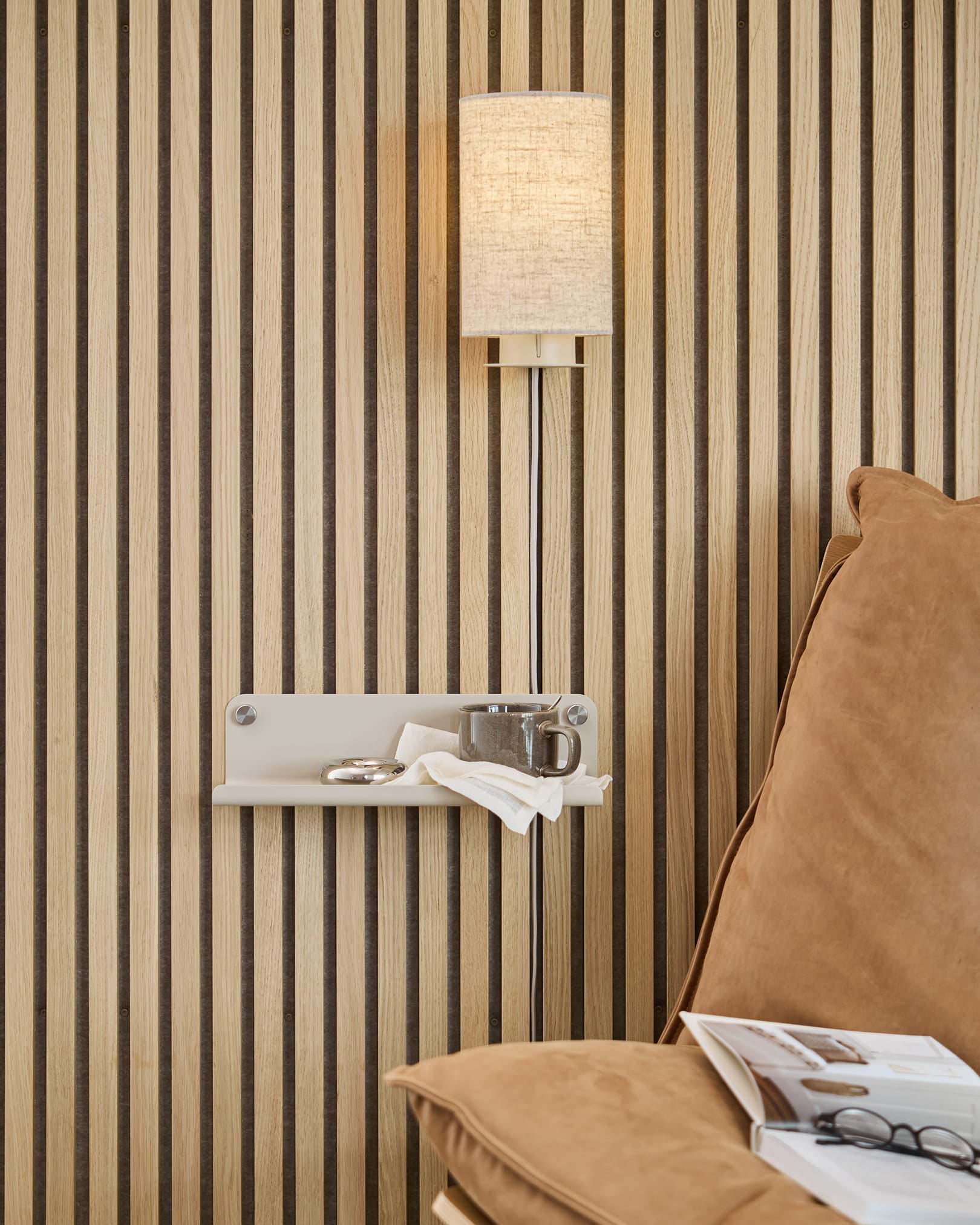 Parete acustica con doghe in legno Akupanel disposte verticalmente, lampada da parete beige, piccola mensola con tazza e tovagliolo, divano color cuoio con una rivista aperta.