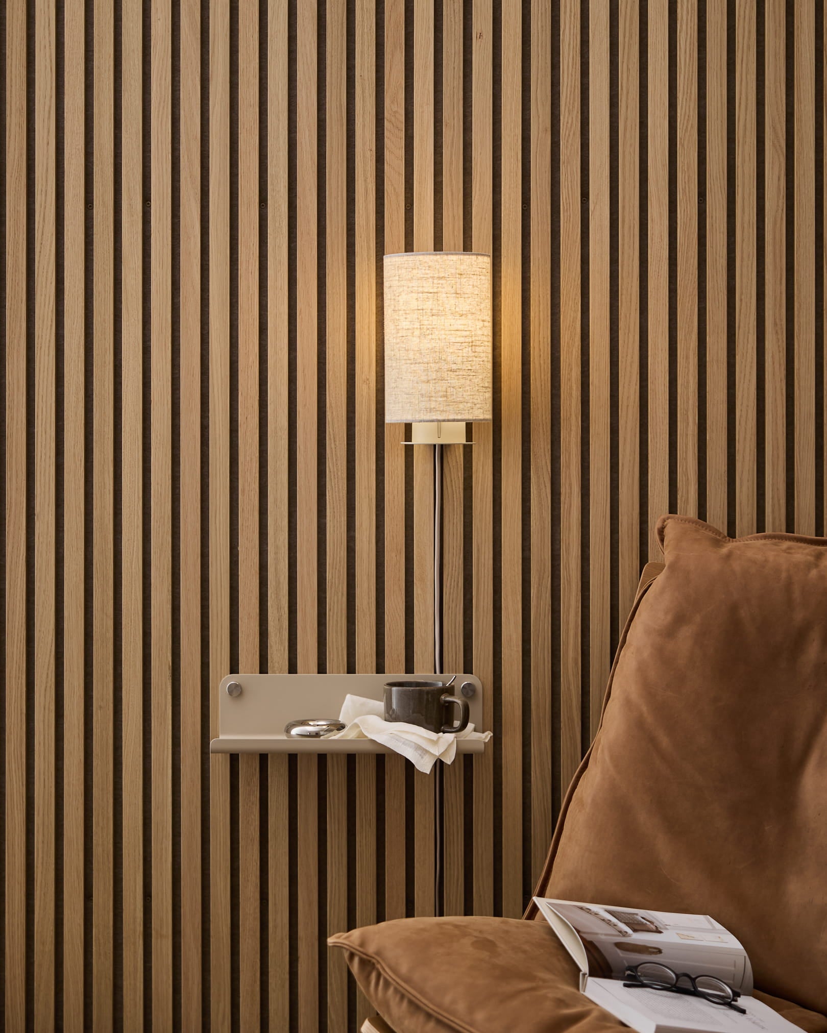 Una Feya Wall Lamp WoodUpp in lino-cotone beige montata sopra una comoda poltrona con tavolino laterale, libro e tazza, sullo sfondo di un rivestimento nordico a doghe in legno Pannello acustico.