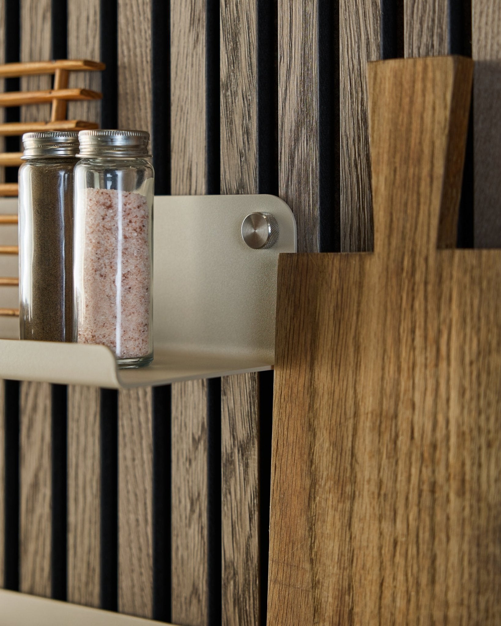 Barattoli di vetro con spezie e un tagliere in legno sono appoggiati sulla mensola larga WoodUpp Norr in beige, 35,9 cm, montata su un pannello acustico a doghe vertical.
