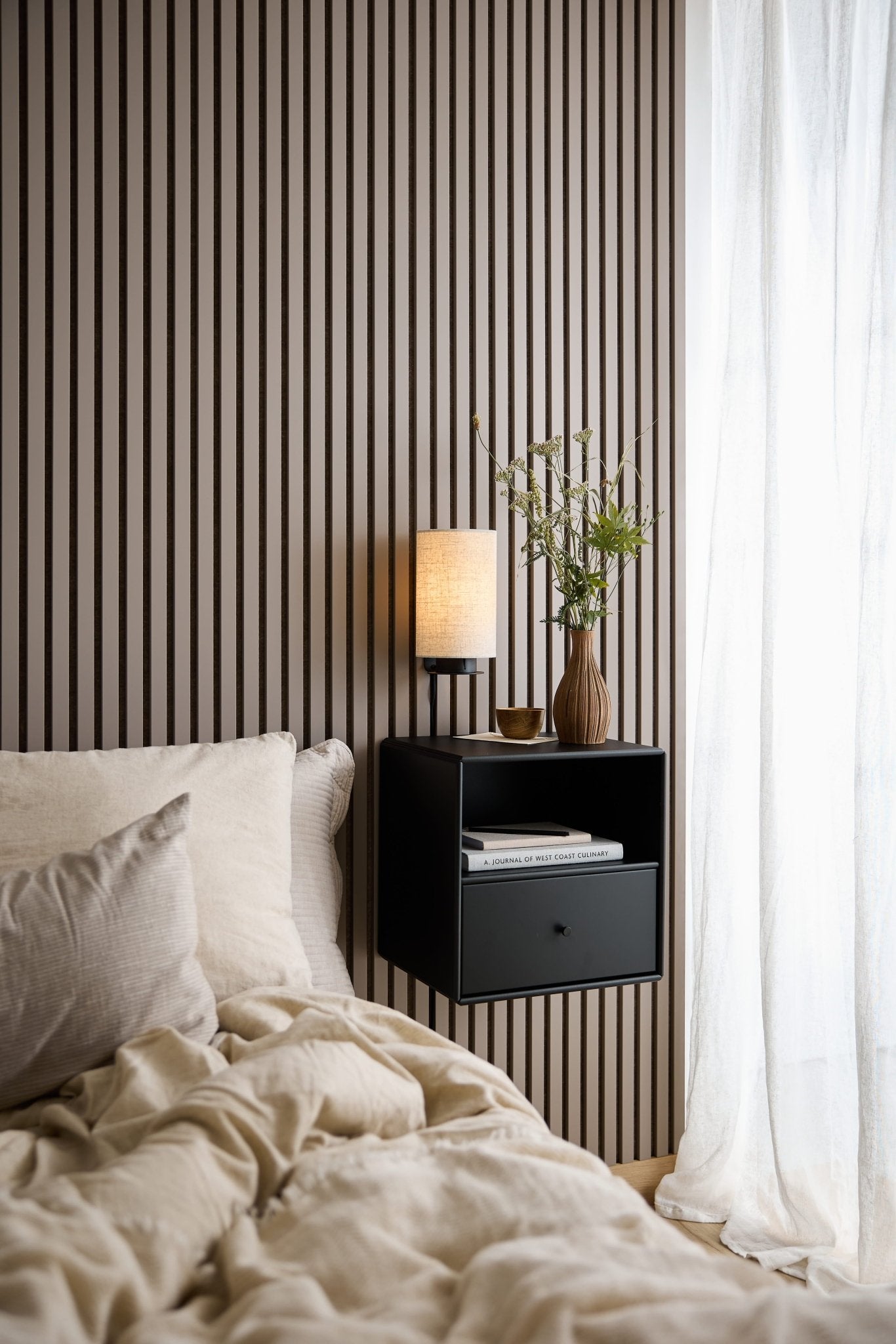 Letto con biancheria da letto beige, cassettiera nera WoodUpp Arka, lampada e vaso vicino a una finestra con tende bianche trasparenti.