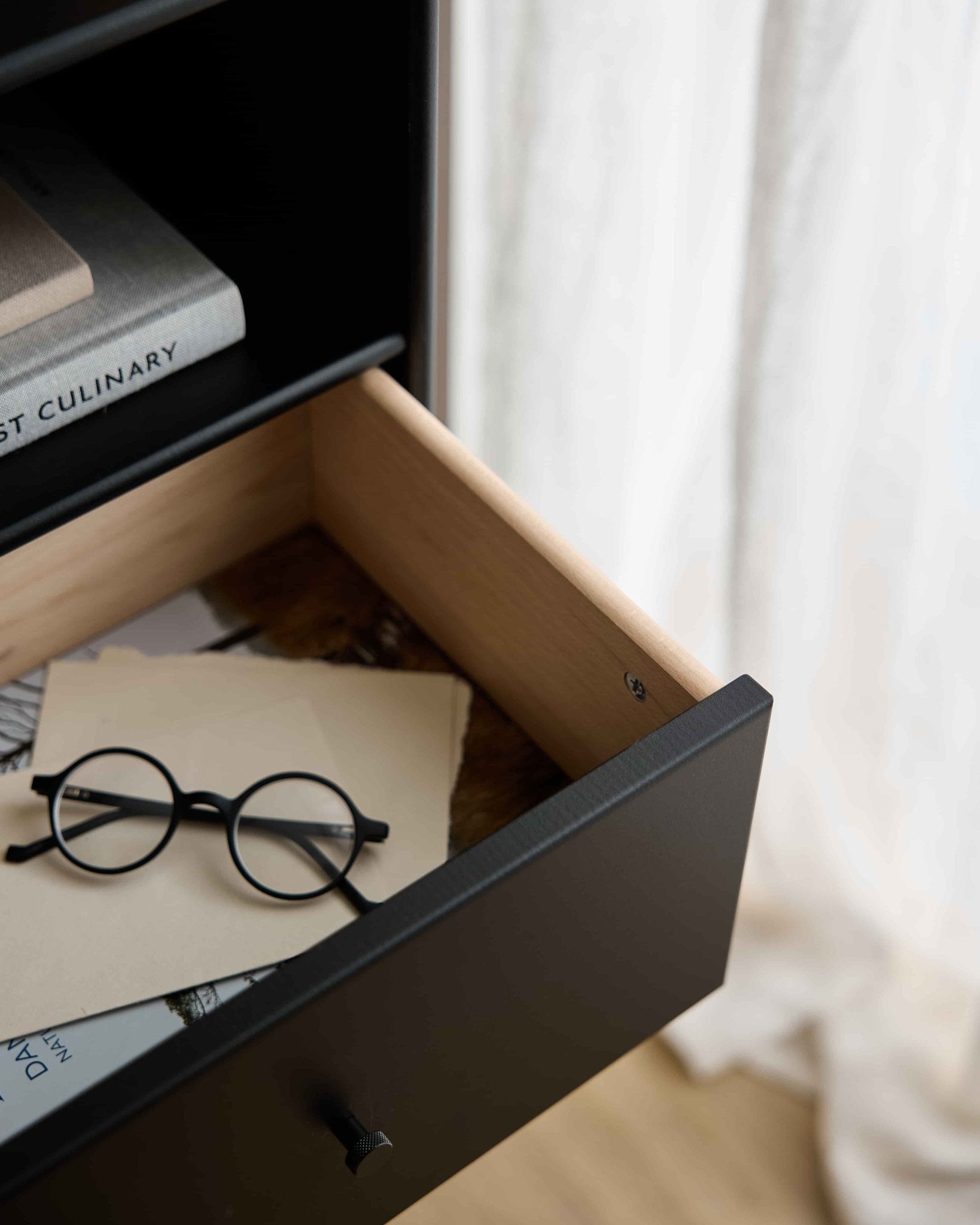 Occhiali rotondi neri e fogli di carta in un cassetto aperto della libreria WoodUpp Arka | Bookcase Drawer / Black, sotto i libri, con tende trasparenti che diffondono delicatamente la luce di fondo.
