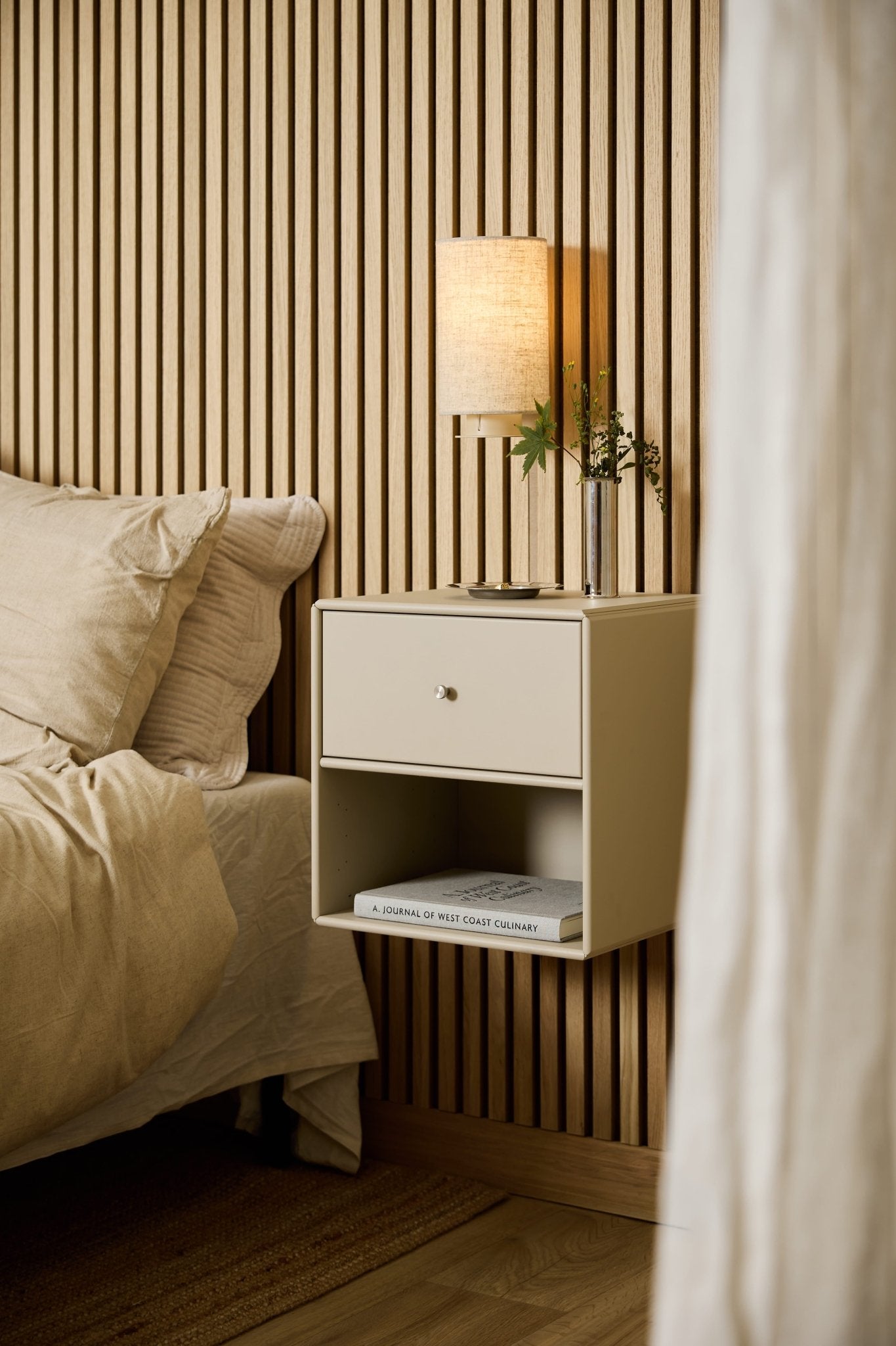 Accanto al letto, una libreria con cassetti WoodUpp Arka di colore beige ospita una lampada, dei libri e un vaso, con alle spalle un pannello acustico in listelli di legno WoodUpp Akupanel.