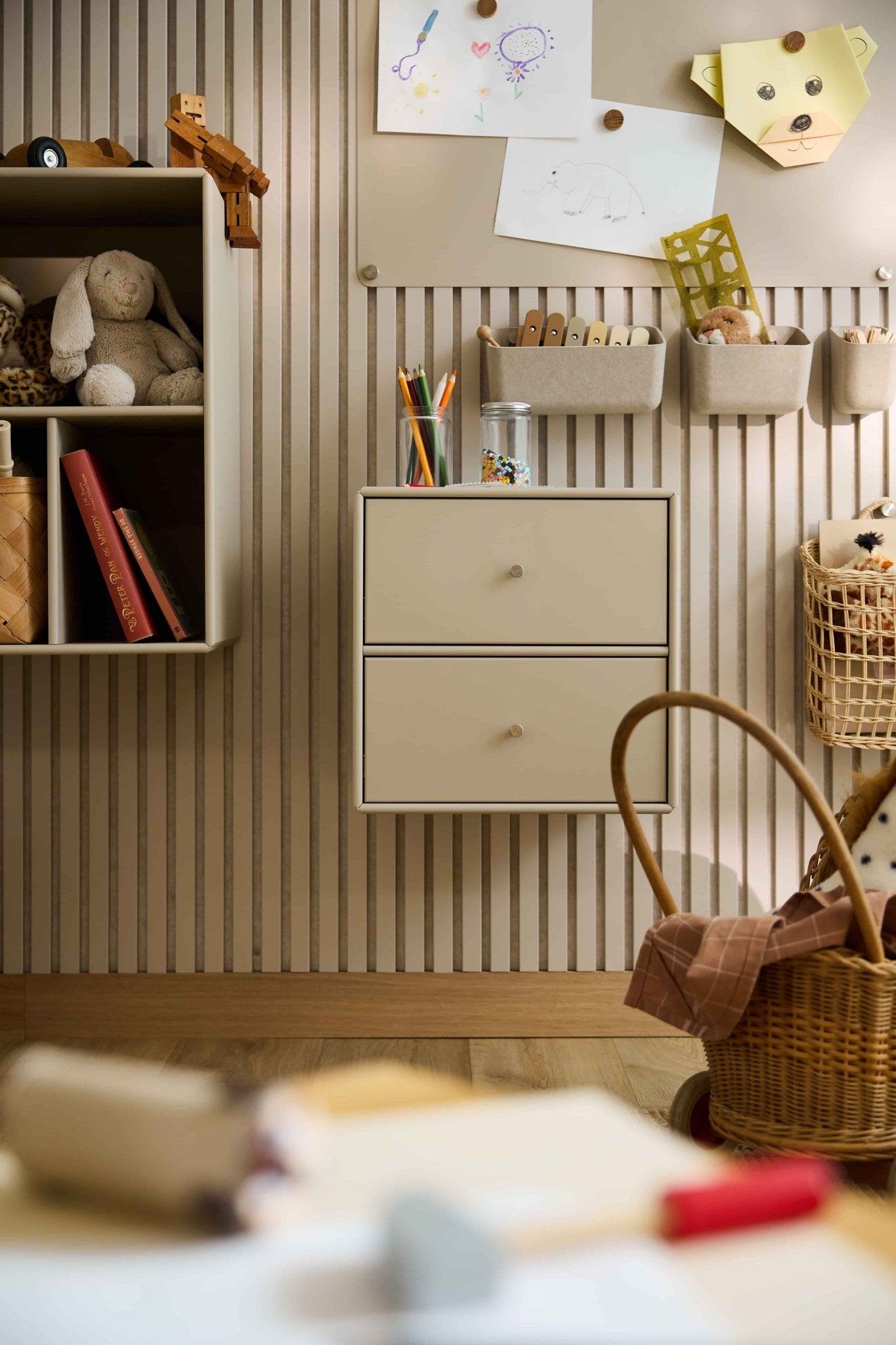 Cameretta con carta da parati a righe, giocattoli, libri, cestini e una libreria con cassetti WoodUpp Arka color beige che offre spazio di archiviazione chiuso.