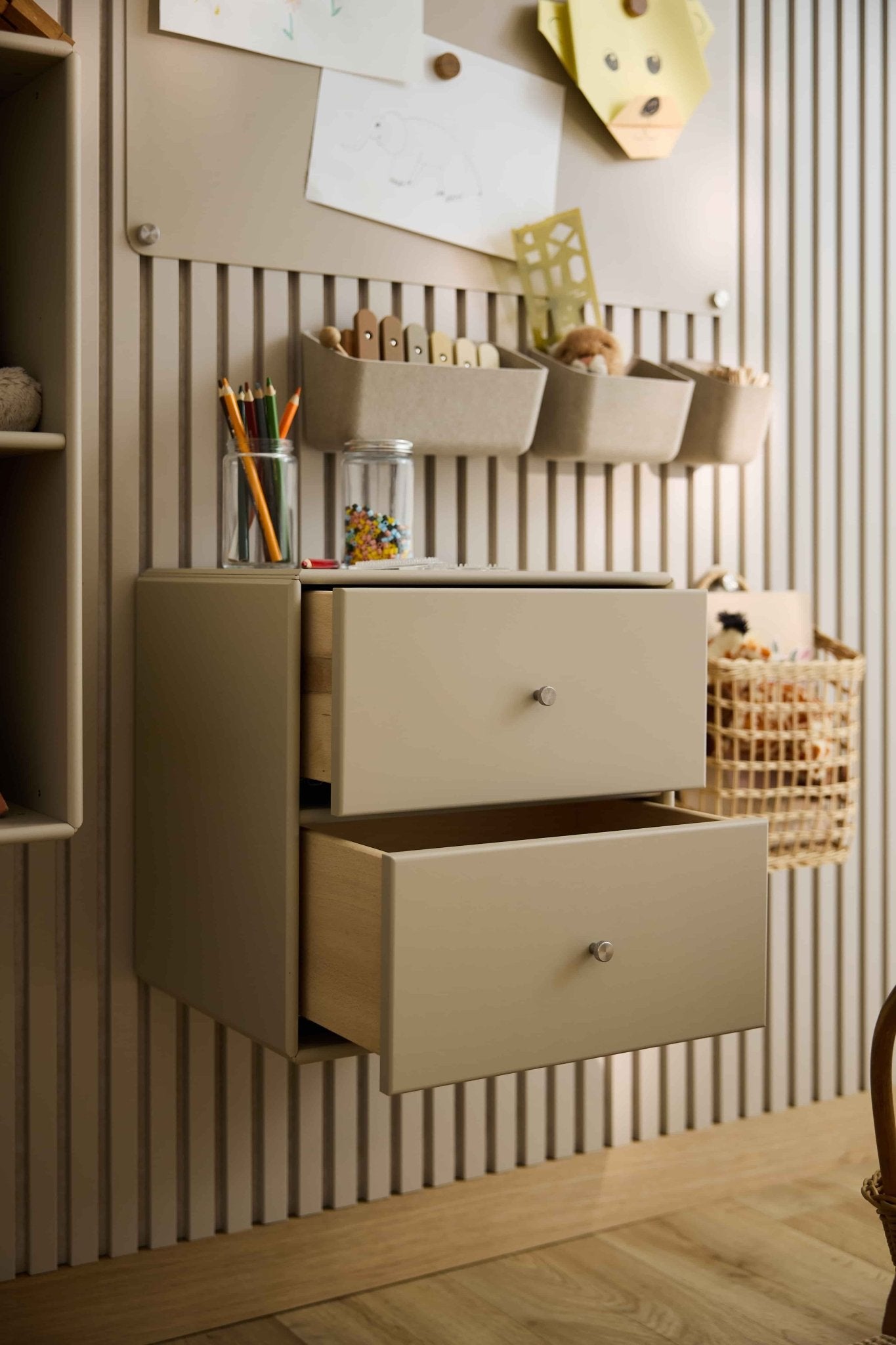 Due cassettiere Arka color beige di WoodUpp, parzialmente aperte, contengono materiale artistico e giocattoli; nelle vicinanze sono collocati contenitori chiusi e cestini.