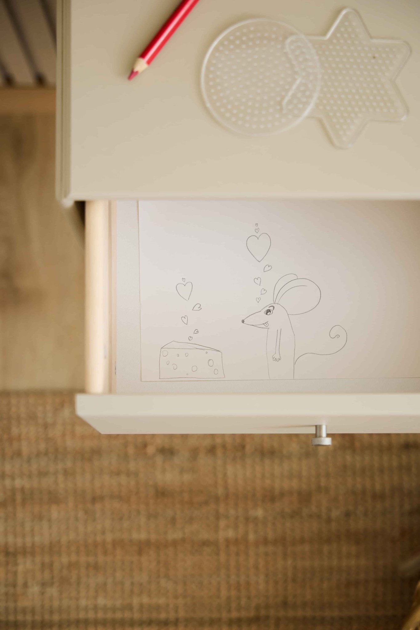 Un topolino e un pezzo di formaggio con motivi a cuori decorano un cassetto della libreria WoodUpp Arka color beige; una matita rossa e dei pannelli forati poggiano sul piano del tavolo abbinato.
