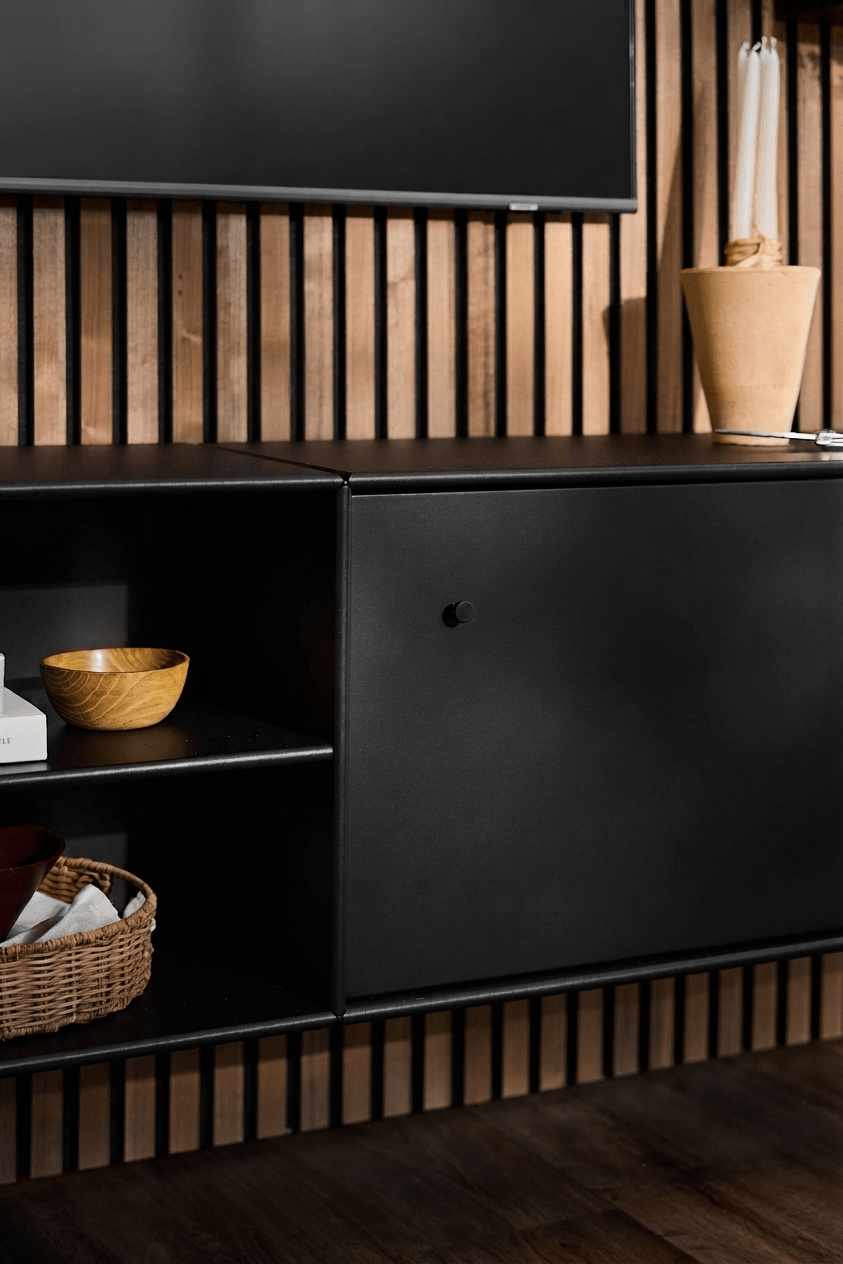 Mobile TV Black WoodUpp Arka con ripiani aperti per libri e cestini, vaso color cuoio, sistema di gestione dei cavi ben studiato e pannello acustico verticale in legno Akupanel come sfondo.