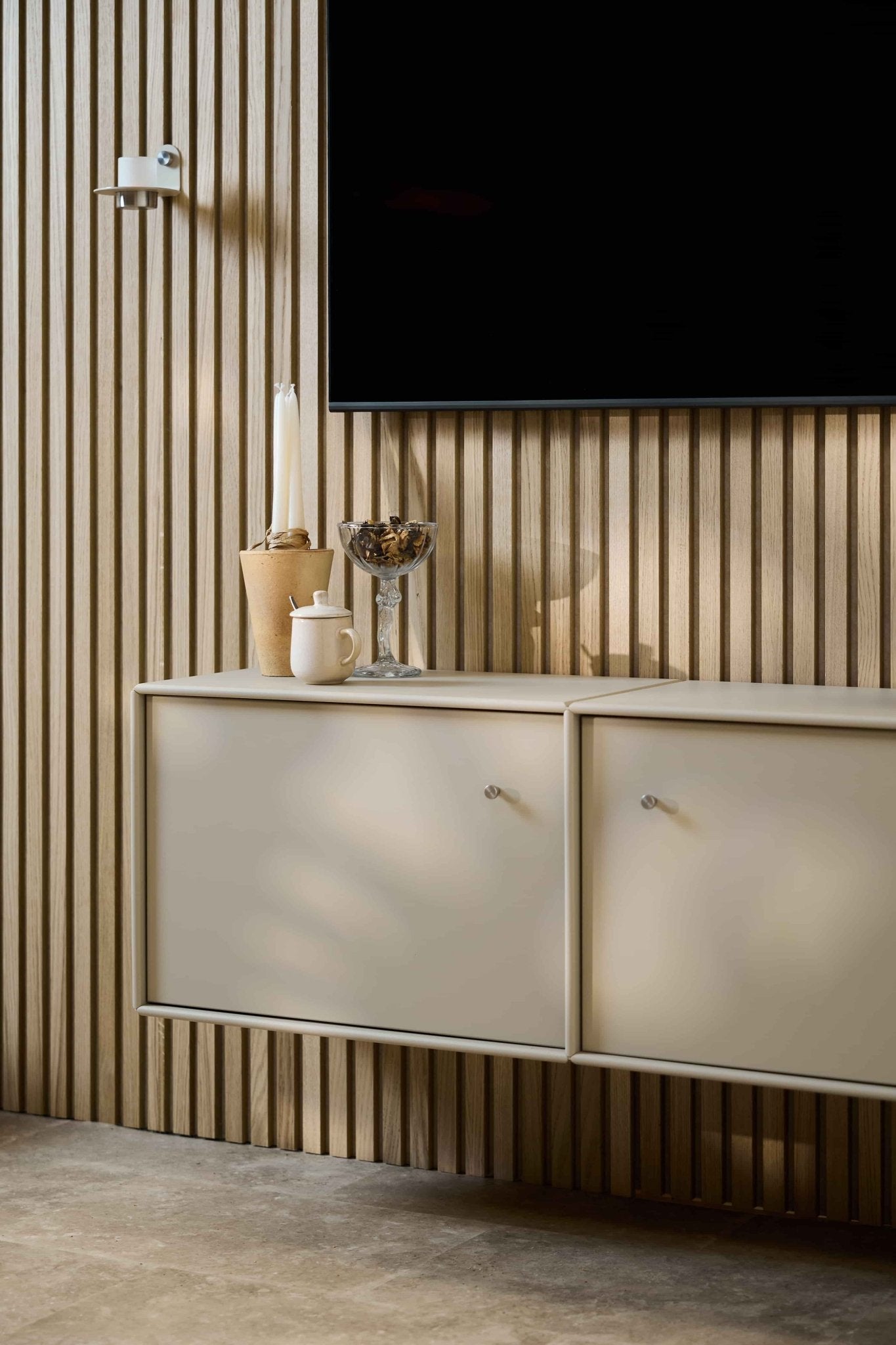 Soggiorno in stile minimalista con mobile TV WoodUpp Arka color beige, parete rivestita con pannelli acustici in legno Akupanel, TV a parete, candele, ciotola di vetro, vaso bianco e cavi ben sistemati.