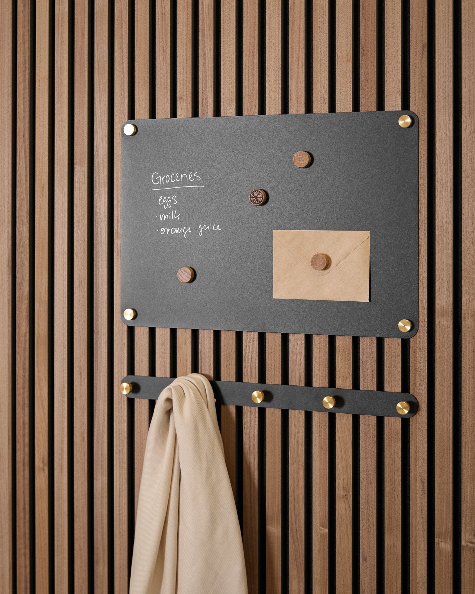 Una lavagna magnetica WoodUpp Norr black small sostiene una lista della spesa, una busta, magneti e un panno beige su ganci, montata su un Akupanel Pannello acustico.