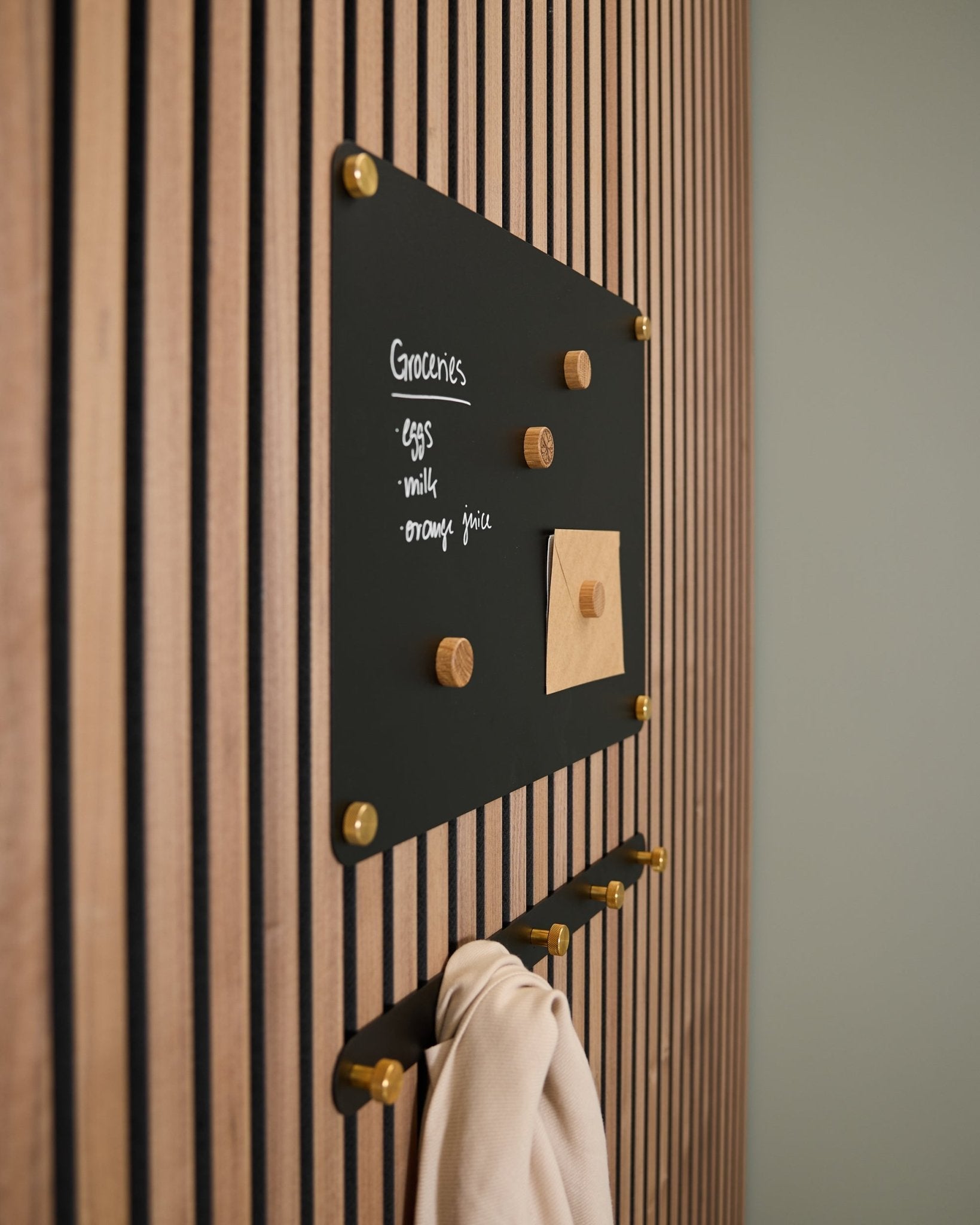 Una lavagna magnetica WoodUpp Norr black small, montata su un pannello acustico in legno Akupanel, espone una lista della spesa e un appunto, con sotto dei ganci che sorreggono un panno leggero.