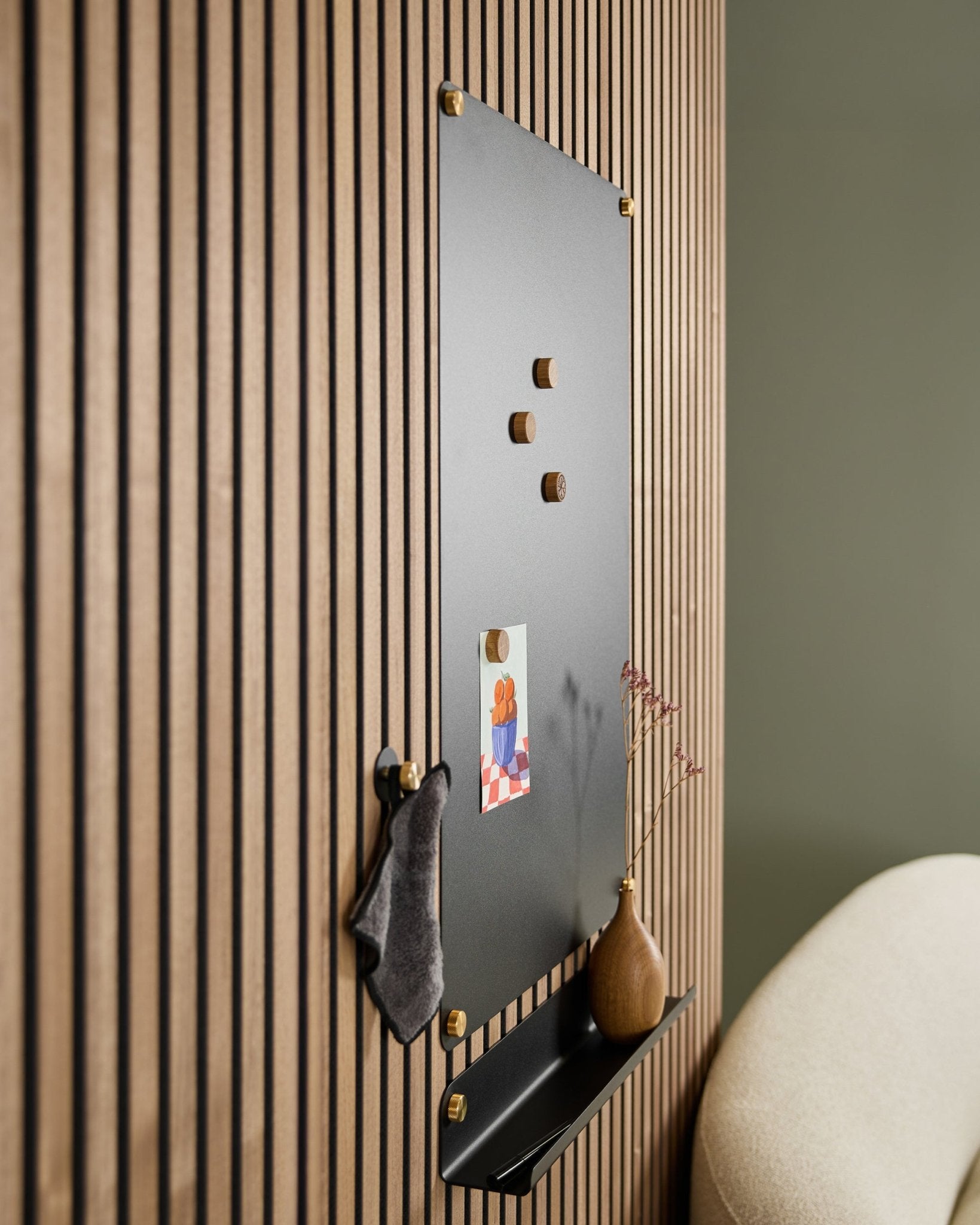 La Norr large black lavagna magnetica acustica WoodUpp con magneti in legno, bacheca, mensola e vaso è montata sulla parete accanto a una sedia beige.