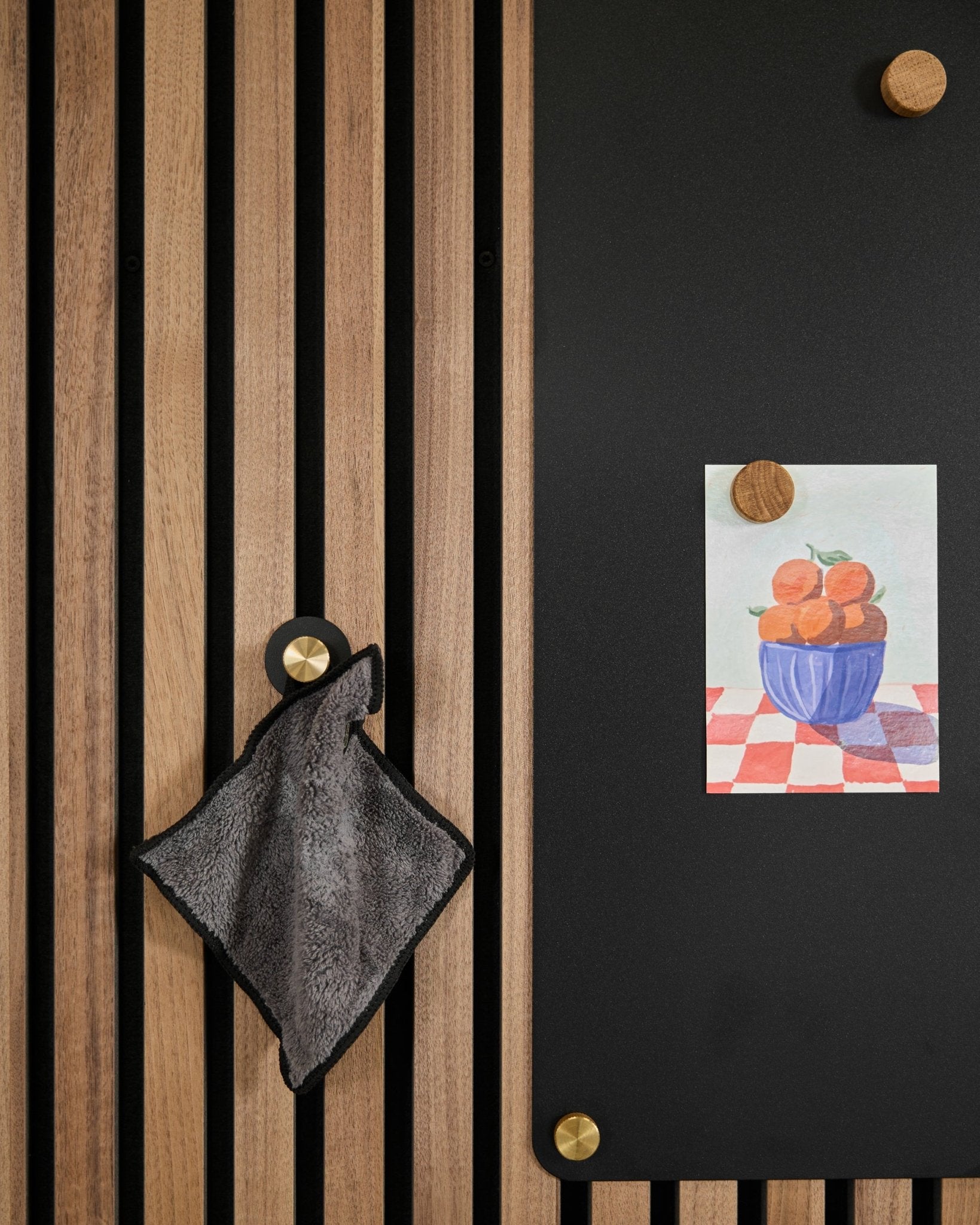 Un asciugamano small è appeso a un pannello acustico a doghe Nordic accanto a una lavagna black che mostra un disegno di arance in una ciotola, fissato con due magneti dorati.