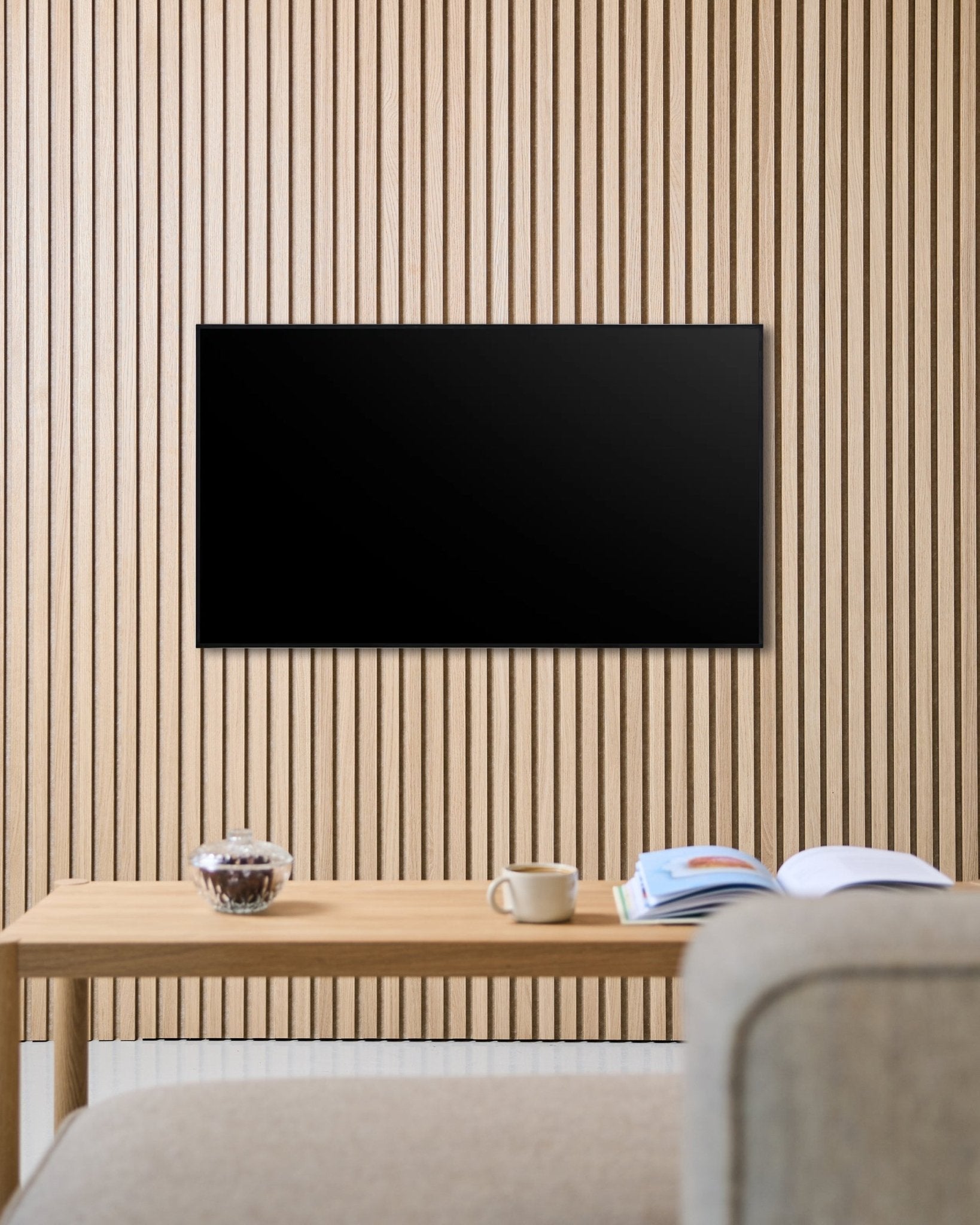 Un supporto TV Fjell (small) di WoodUpp fissa uno schermo piatto a una parete acustica a doghe in legno Nordic Akupanel, sopra un tavolo con tazza, ciotola e una rivista aperta accanto a un divano beige.