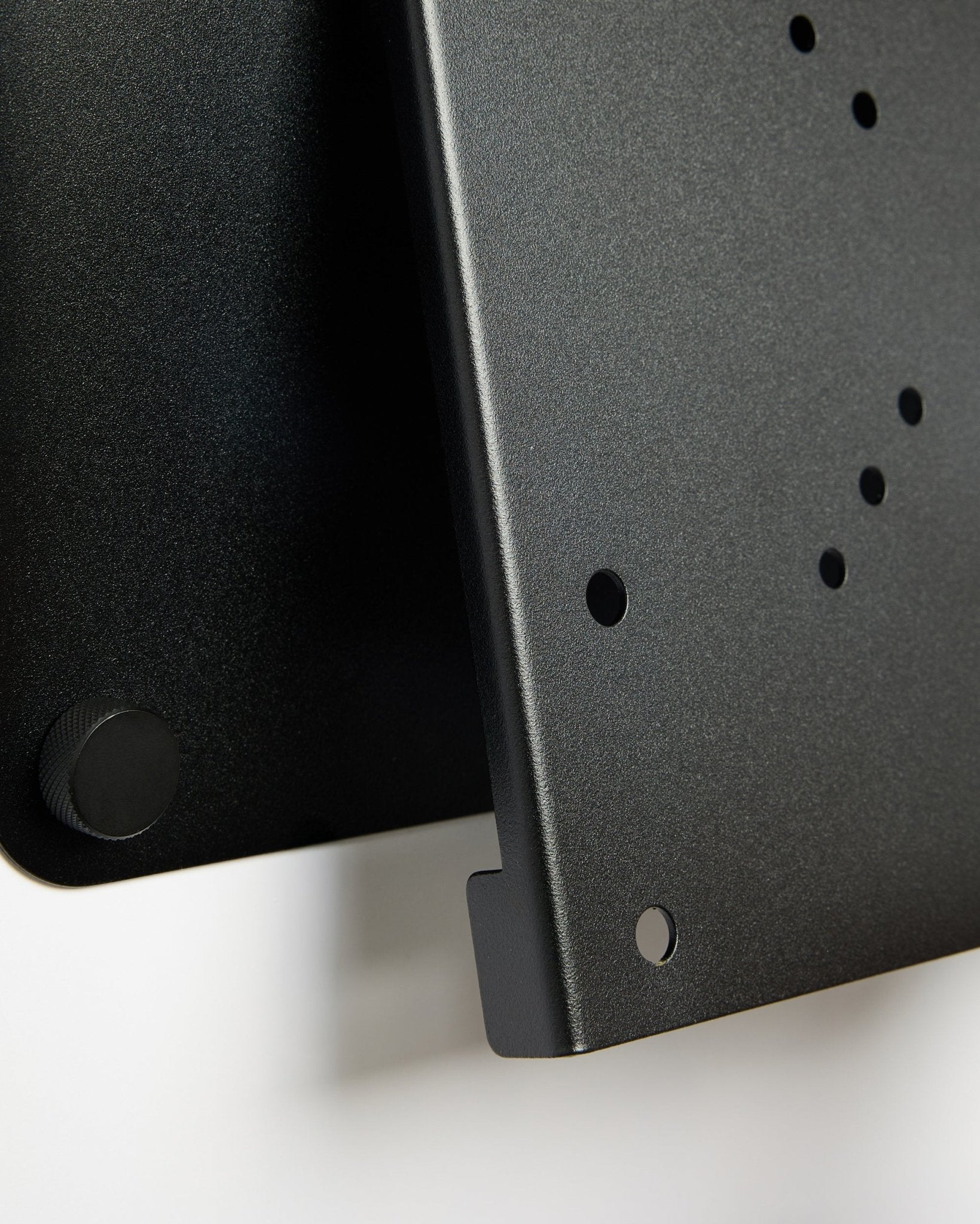 Primo piano del supporto TV Fjell (small) di WoodUpp, una staffa in metallo black con fori circolari e manopola di regolazione, mostrata su sfondo bianco.
