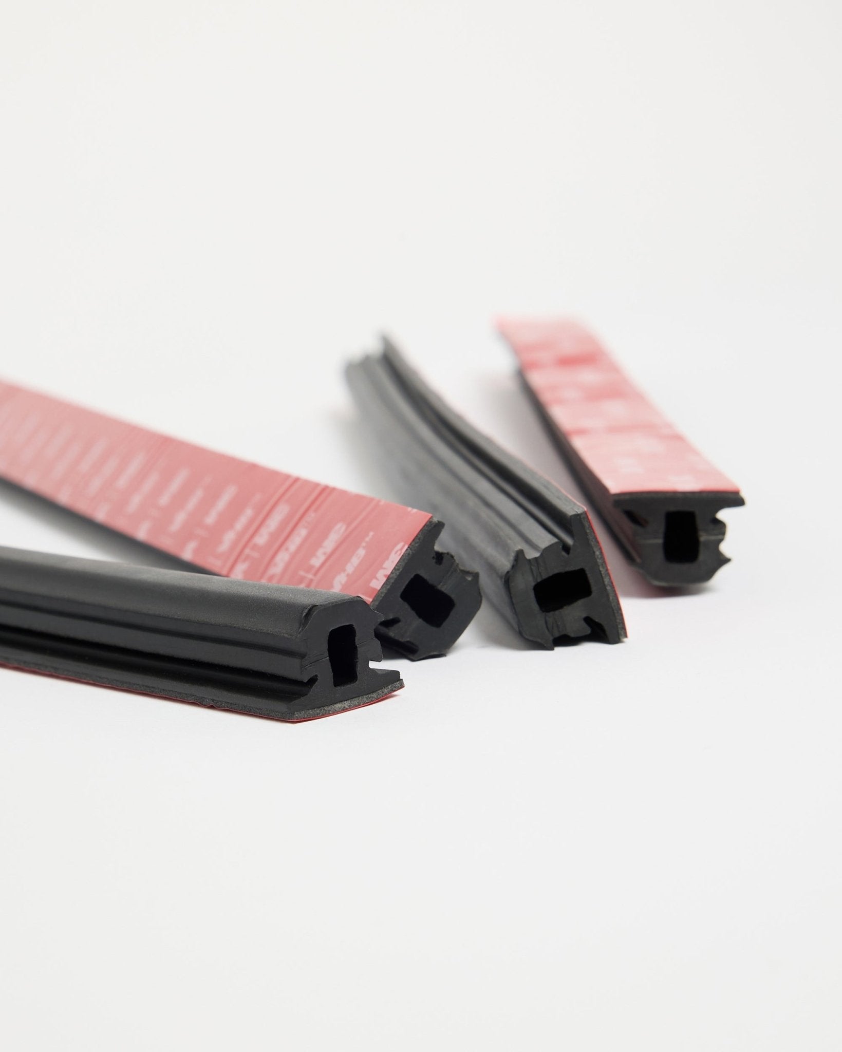 Quattro strisce in gomma Pop-on Profile WoodUpp Dupps black con adesivo rosso, pensate per un’installazione Akupanel, disposte su una superficie bianca in diverse direzioni.