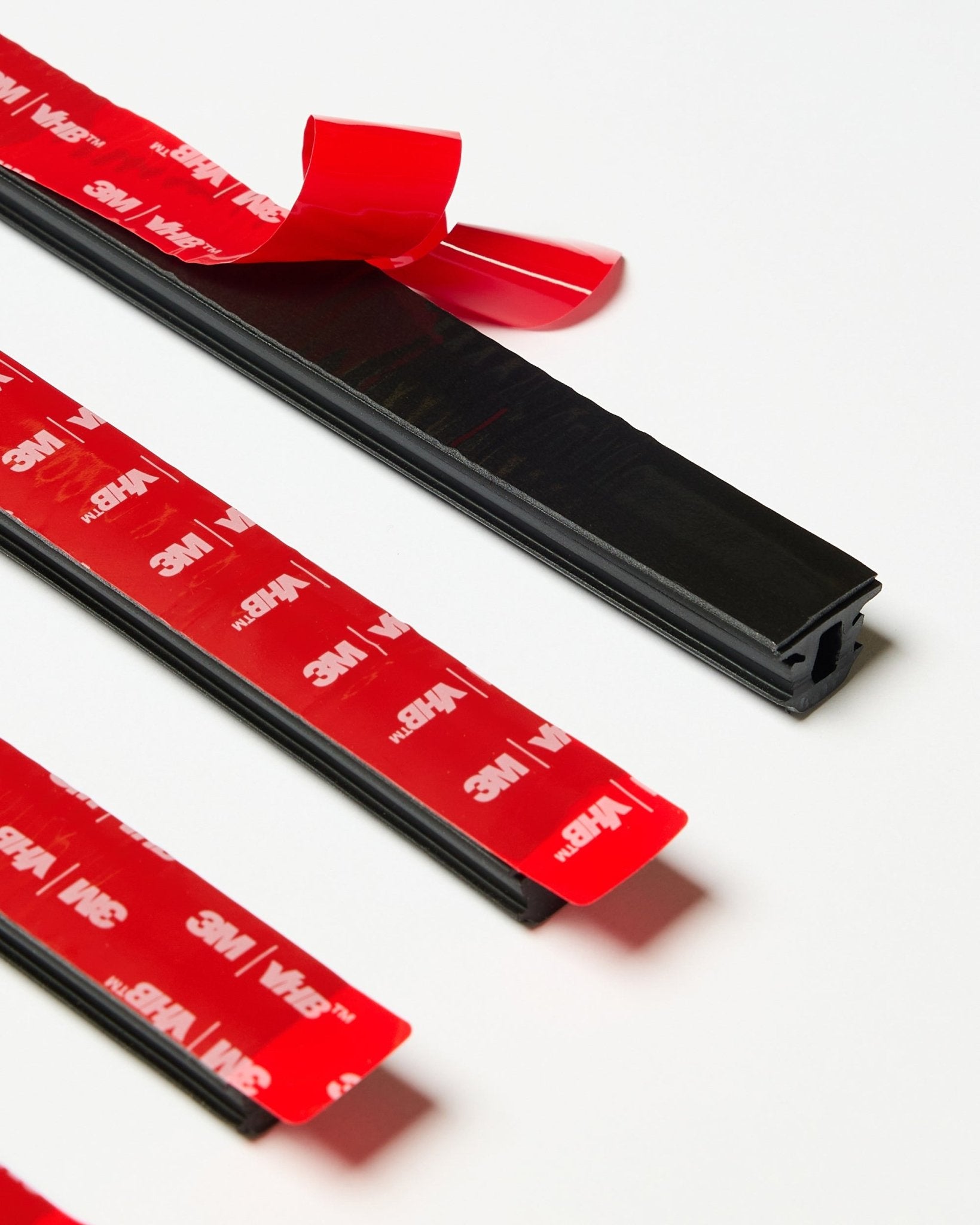 Set di quattro strisce WoodUpp CREATE Pop-on Profile black con supporto adesivo 3M rosso visibile, una striscia parzialmente staccata, presentate su una superficie bianca.