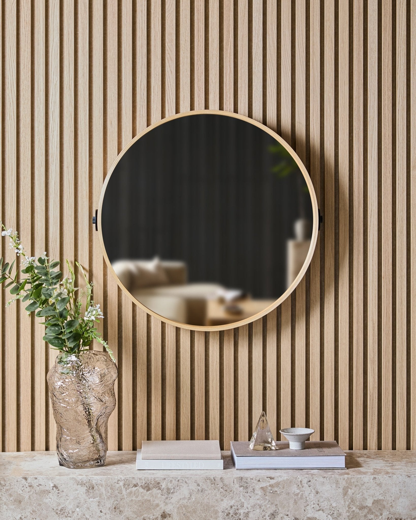 Il Linn | Mirror in Natural Oak di WoodUpp è montato su una slatwall nordica Pannello acustico sopra una mensola in pietra con un vaso in vetro, libri e oggetti decorativi.